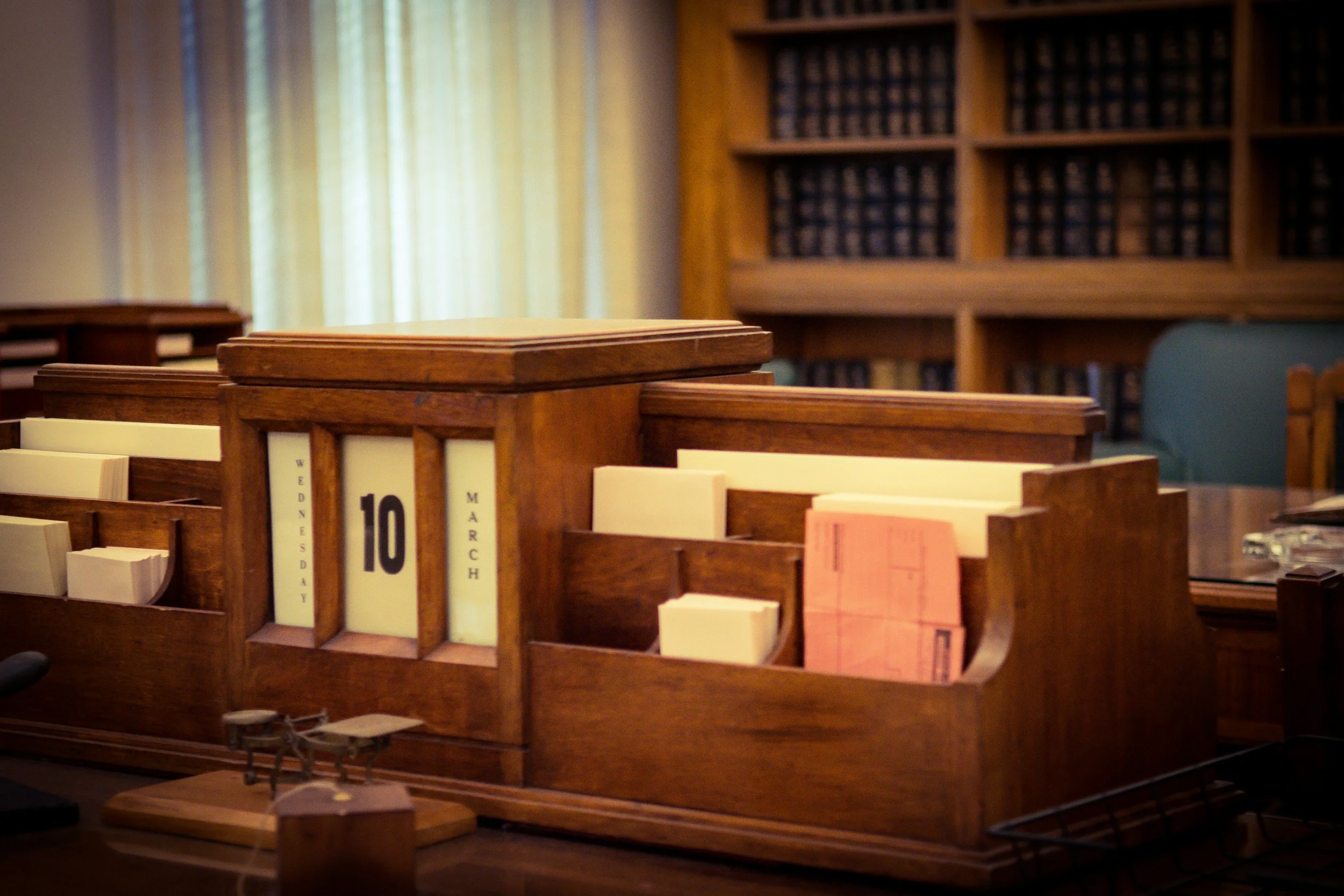 A wooden desk calendar displaying March 10, Wednesday, with files and papers in an office or library setting, with bookshelves in the background.
