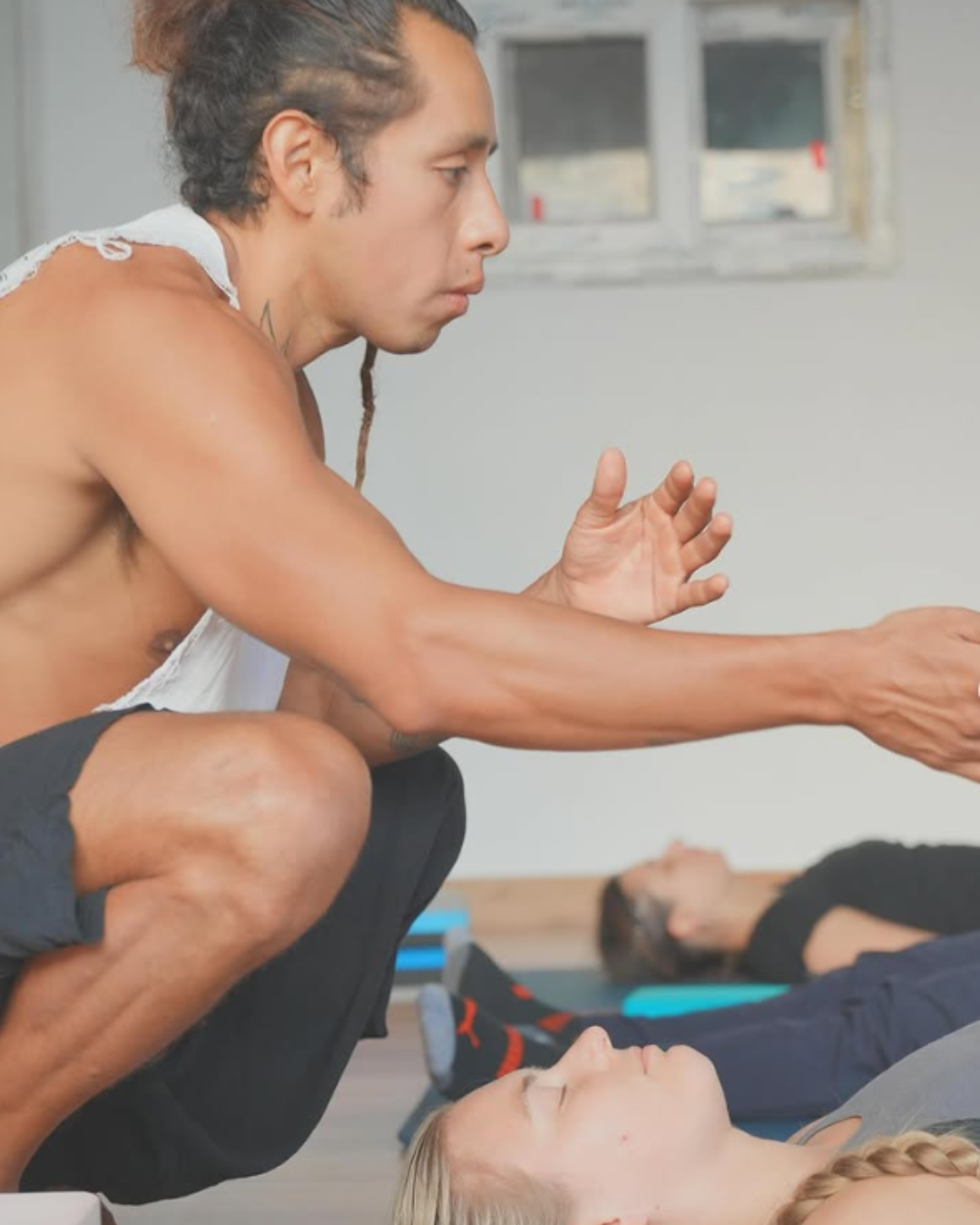 A yoga or fitness instructor helps a woman with her exercise, with another participant practicing in the background.