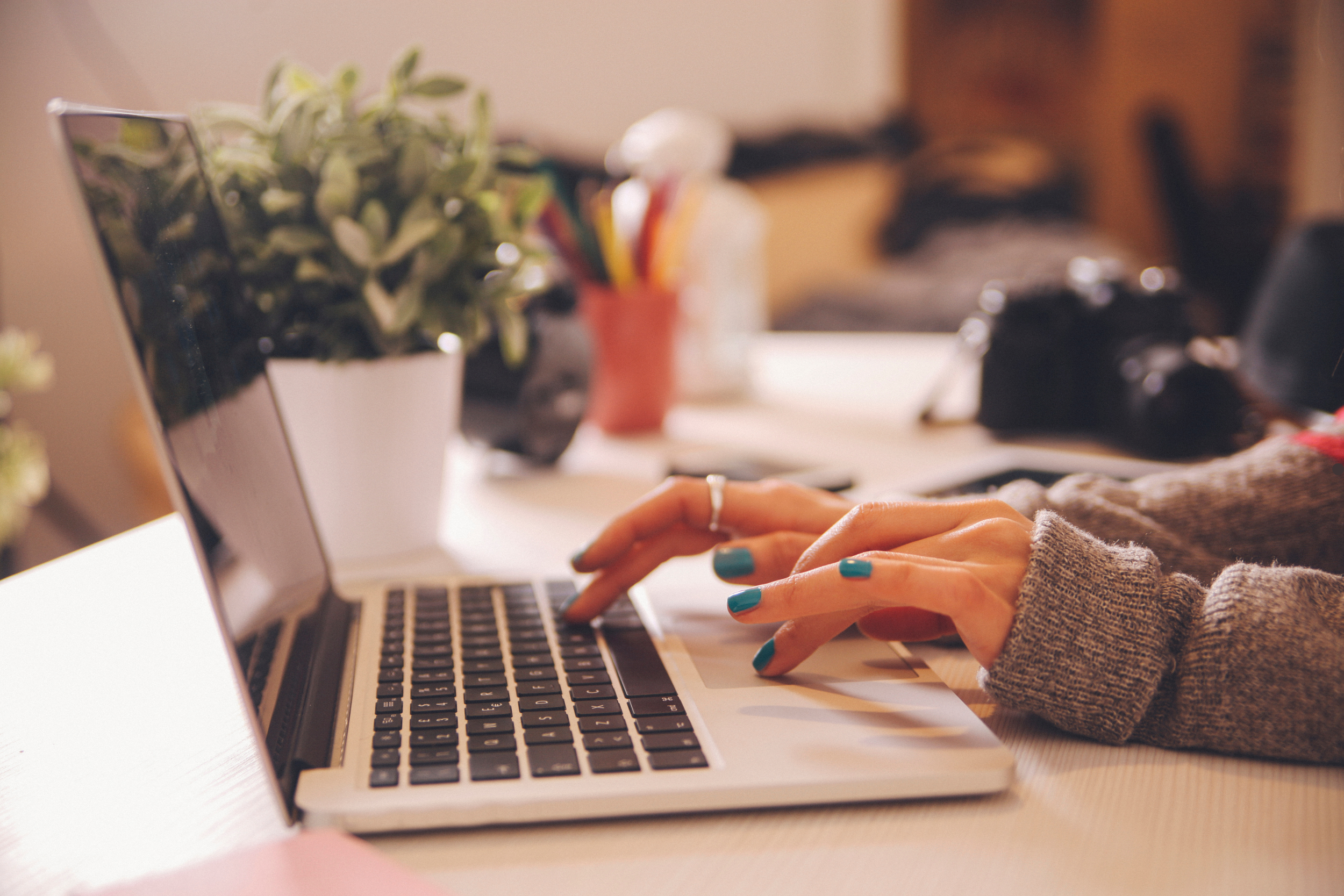 Person typing on a laptop keyboard at a desk, with a potted plant and office supplies in the background.