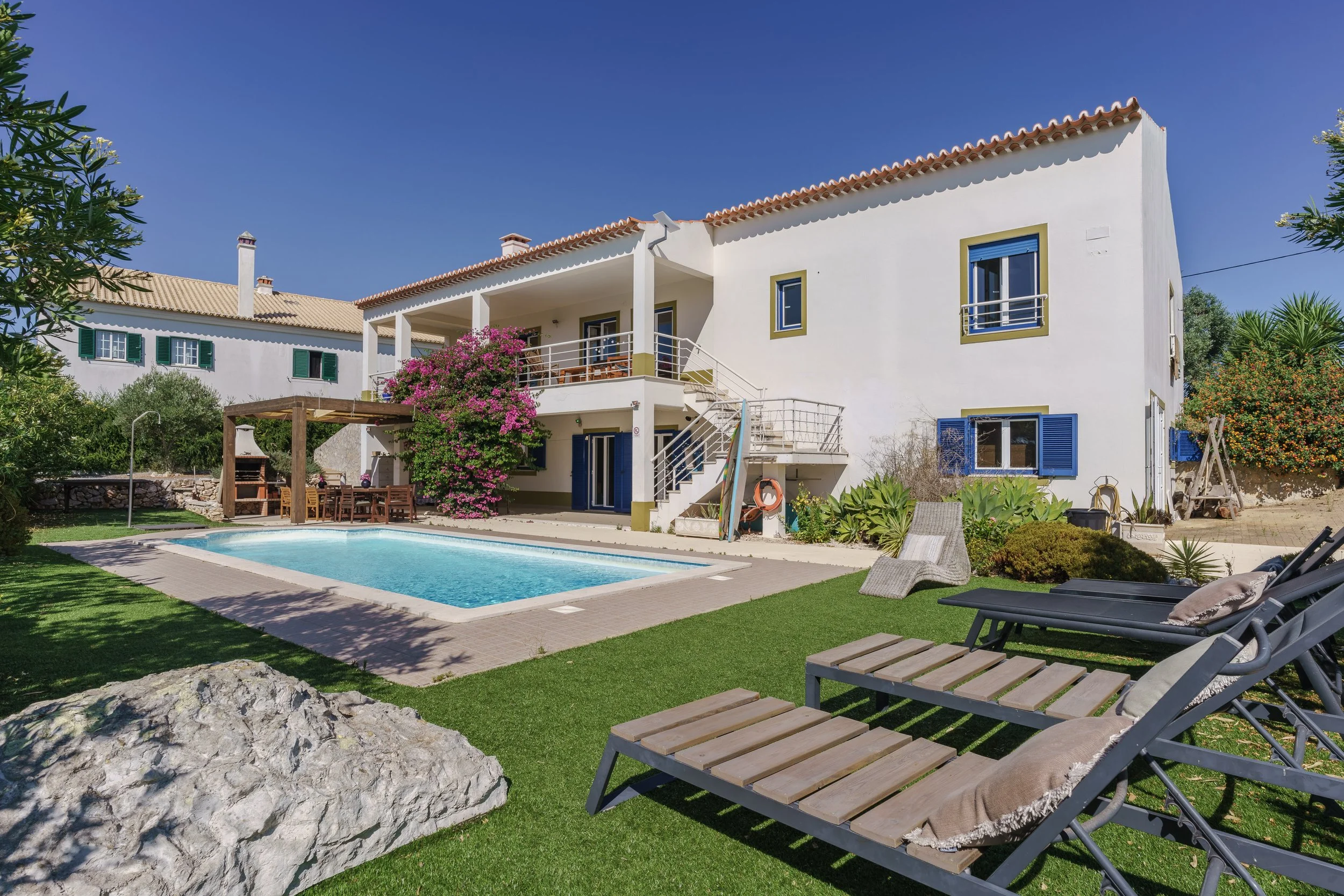 A two-story white house with blue shutters, a swimming pool in the backyard, a wooden pergola with outdoor dining, lounge chairs, and lush greenery under a clear blue sky.