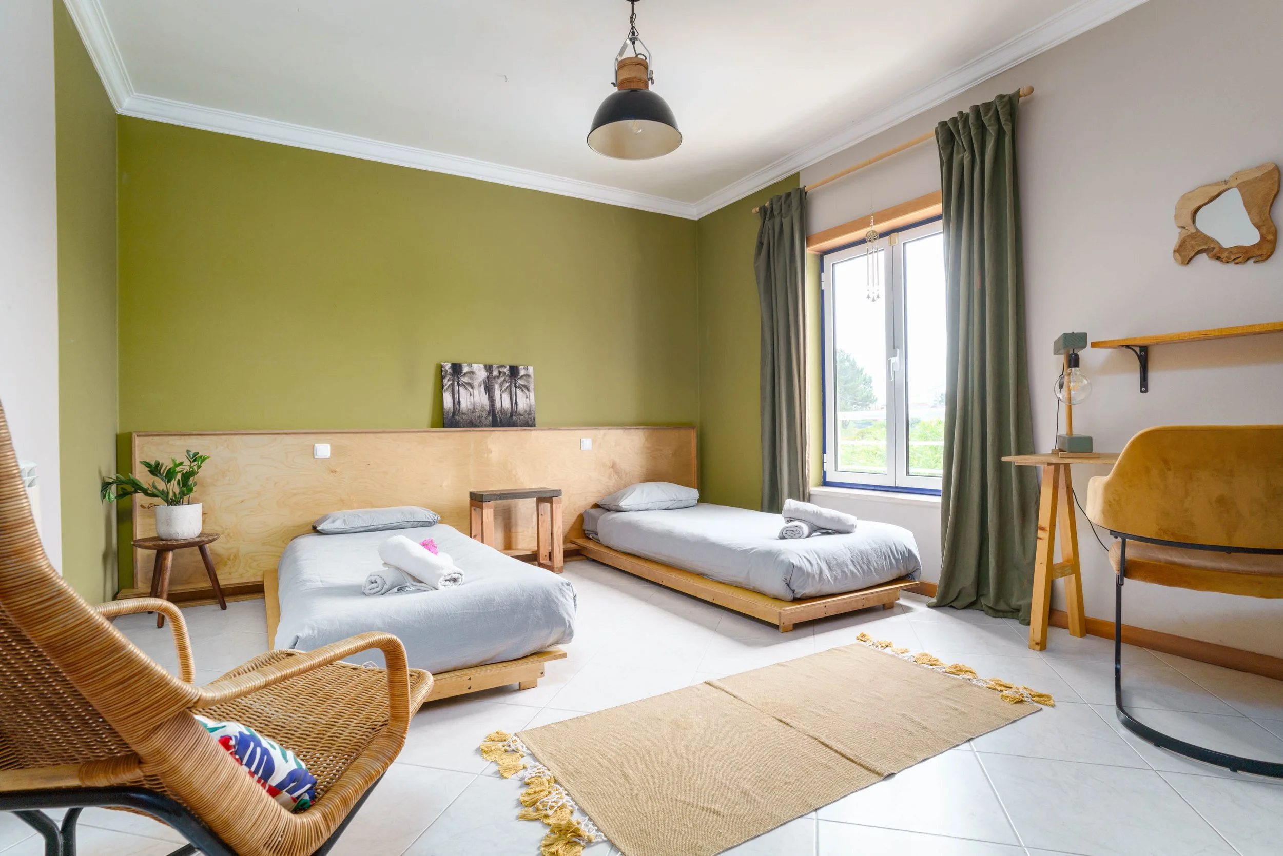Room with two beds, a wicker chair, a desk with a lamp, a window with green curtains, and a green accent wall.