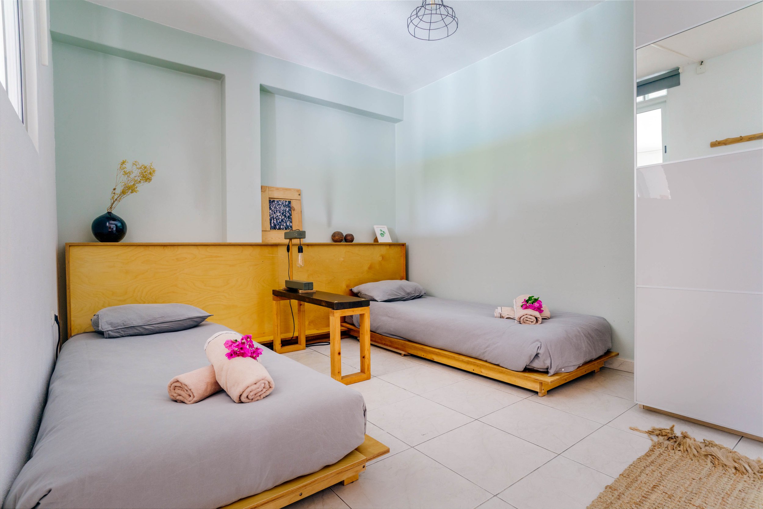 Two single beds with gray bedding and rolled towels topped with pink flowers in a bright bedroom with white walls and a tiled floor, decorated with a black vase with yellow flowers, framed artwork, and a bedside lamp.