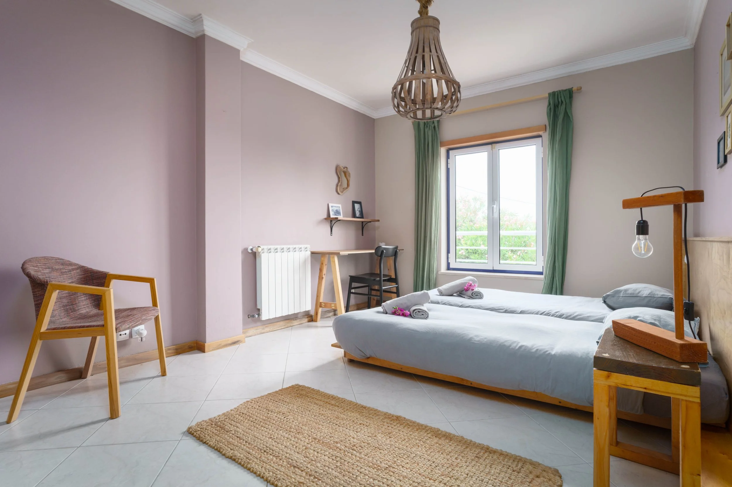 A bedroom with two beds, a window with green curtains, a small desk with a chair, a bedside table with a wooden lamp, a chair, and a beige rug on white tiled floor.