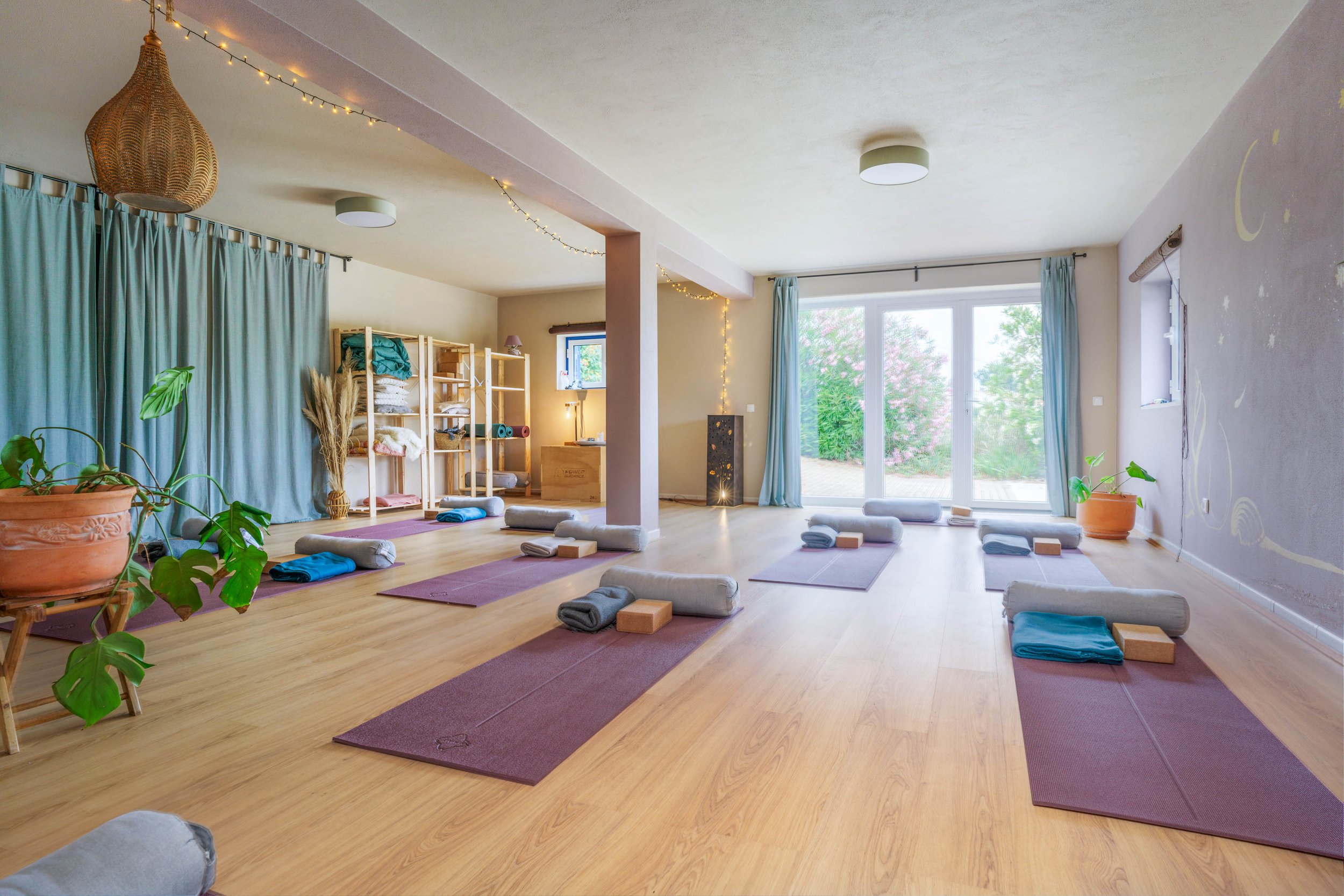 Yoga studio with purple mats, rolled towels, blocks, and cushions on a wooden floor. Large windows with blue curtains, potted plants, fairy lights, and wall decorations.