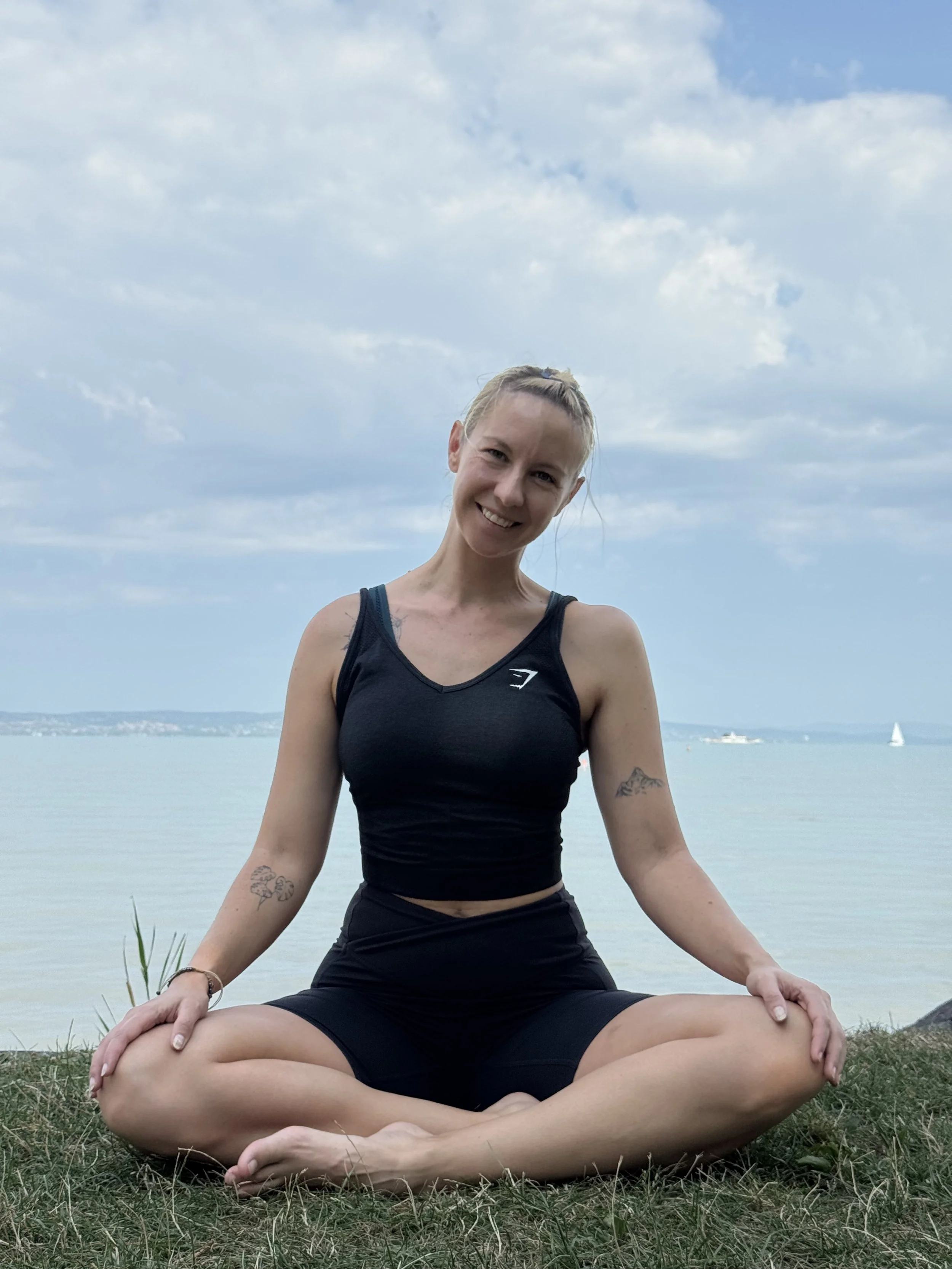 A woman sitting cross-legged in a meditation pose on the grass by the water, smiling at the camera with sailboats and a cloudy sky in the background.