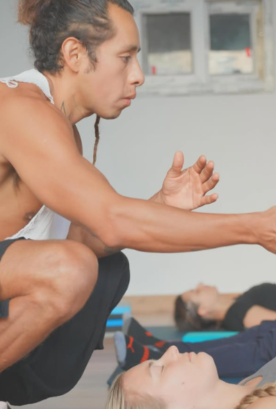 A young man with long curly hair and a sleeveless shirt is in a yoga or meditation class, sitting on the floor with his knees bent. He appears to be guiding or instructing a woman lying on the floor with her eyes closed, who is participating in the session. There is a woman lying on a mat in the background.