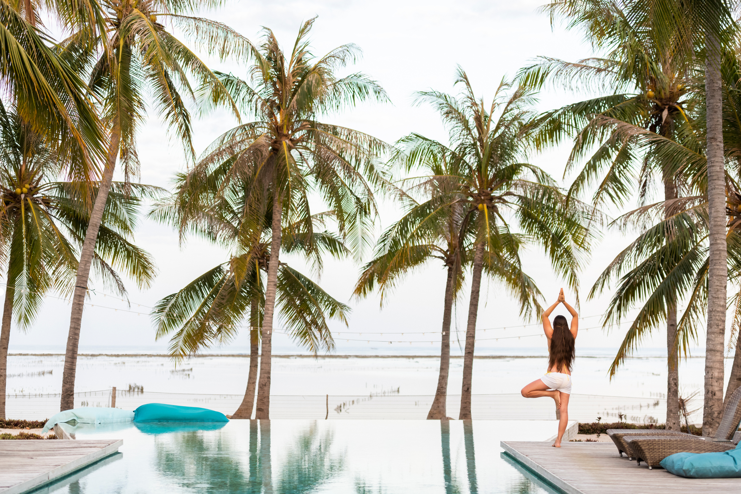 Woman practicing yoga in a tree pose near an infinity pool with palm trees and a body of water in the background at a tropical beach resort.