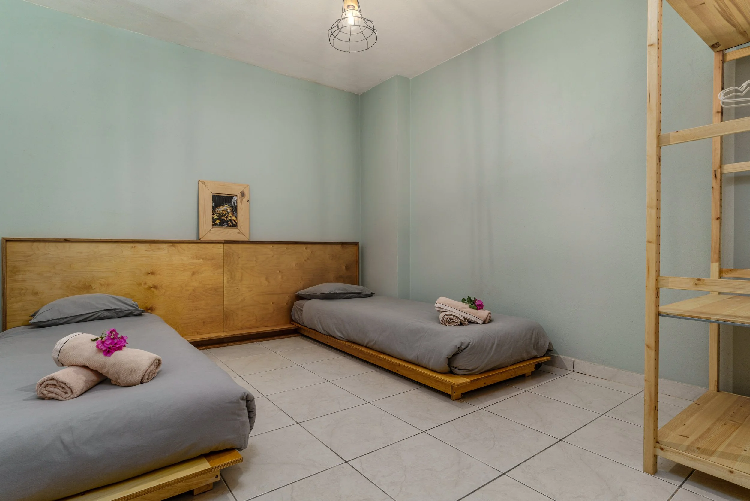 A minimalist bedroom with two beds, each with gray bedding and flowers, against a pastel green wall, with a wooden headboard, framed picture, and a wooden shelf unit.