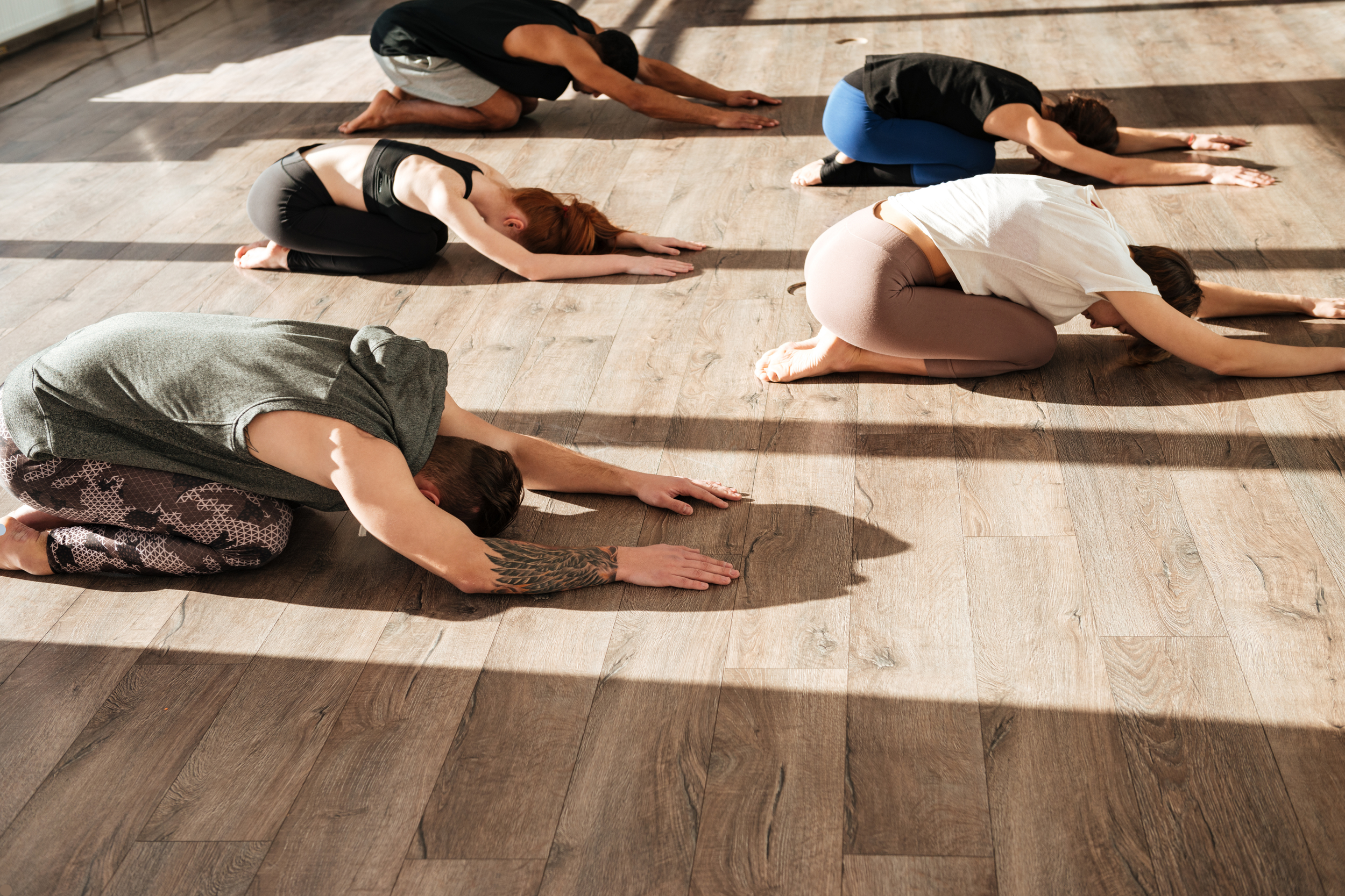 Group of people practicing yoga indoors on a wood floor, in child's pose with knees and head touching the ground, arms extended forward.