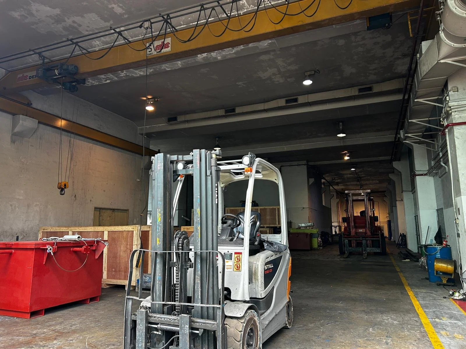 An indoor industrial or warehouse space with a forklift in the foreground, large red container, crates, and various equipment and tools.