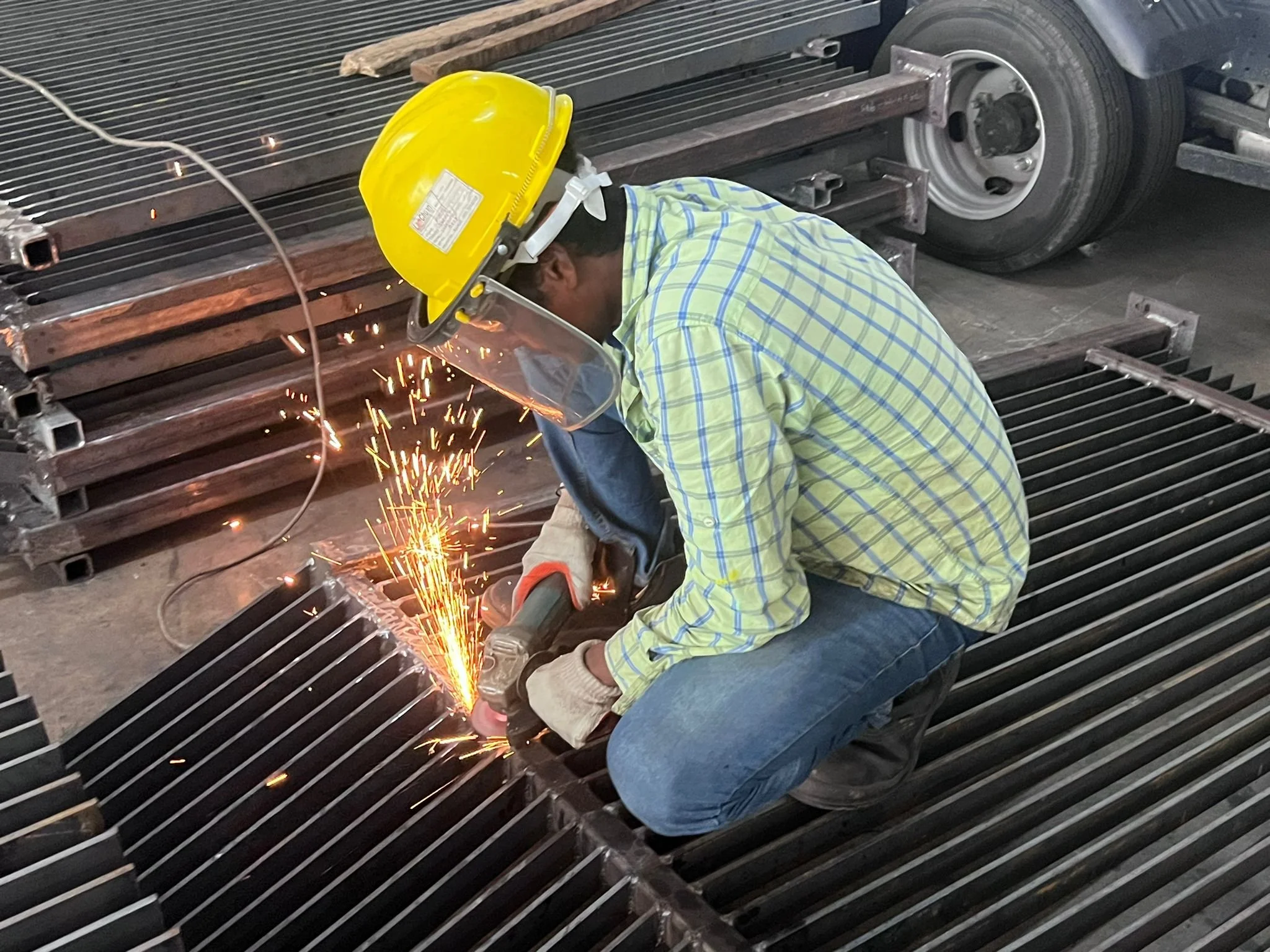 A worker welding metal grates, wearing a yellow safety helmet, face shield, gloves, and a yellow plaid shirt, surrounded by stacks of similar metal grates in an industrial setting.