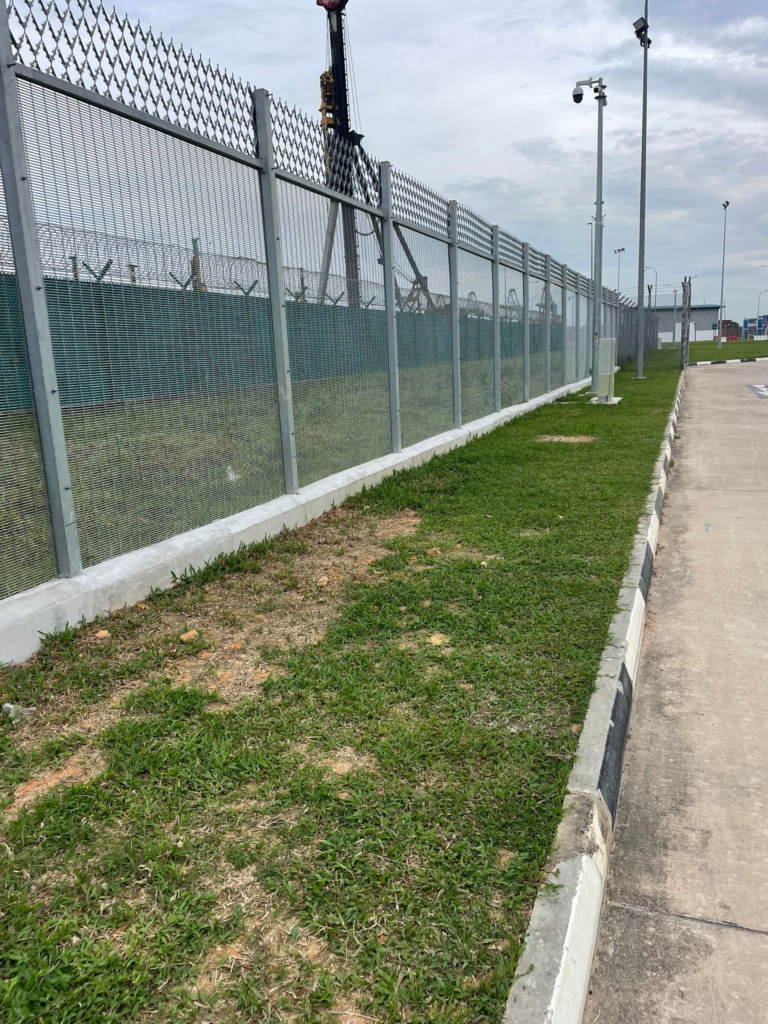 A tall metal security fence with barbed wire on top, running alongside a grassy area and a concrete sidewalk. Behind the fence, there is a crane and some industrial structures, with cloudy sky overhead.