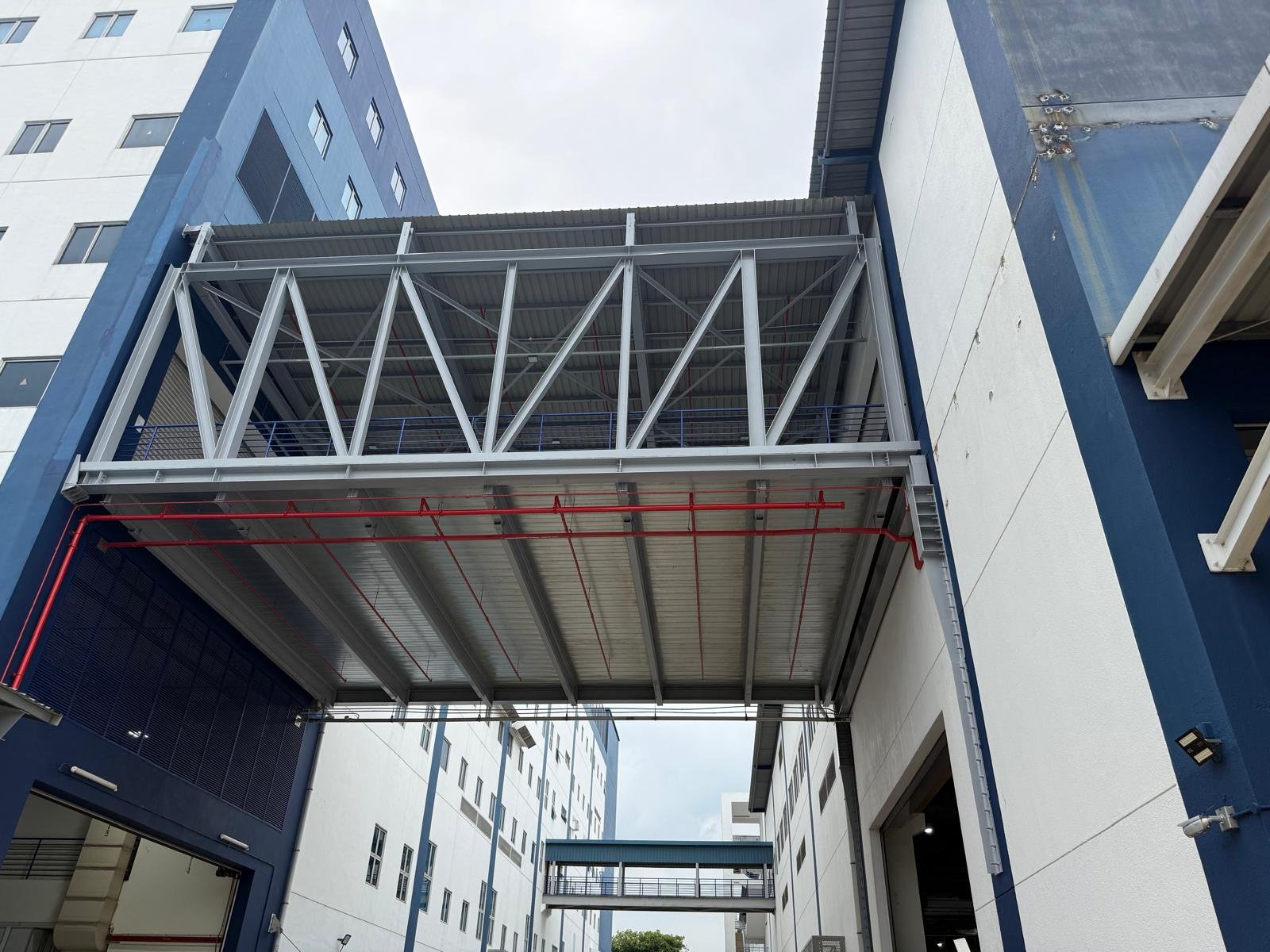 View of a multi-story building with an external metal and glass balcony or walkway under construction, featuring scaffolding and red piping, in an urban setting.
