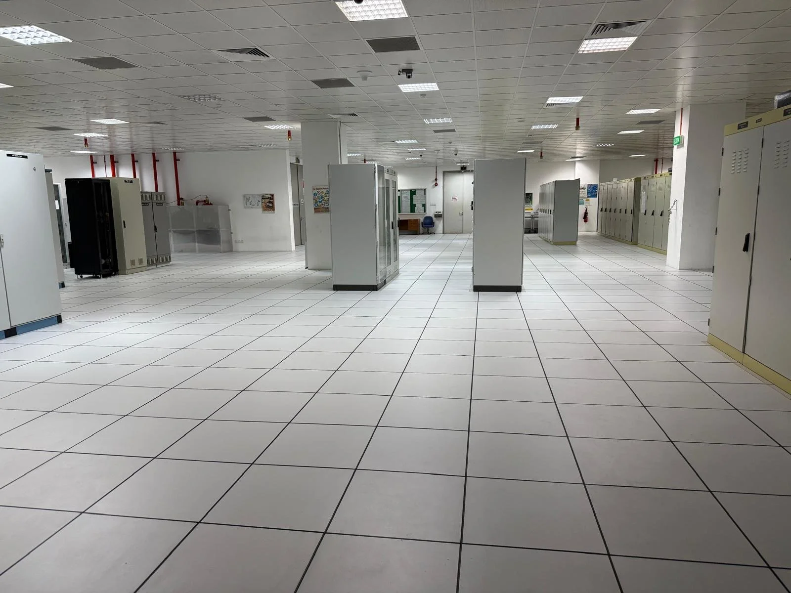 Empty office space with white tiled floors, white walls, and ceiling, lined with metal lockers, cabinets, and some chairs in the background.