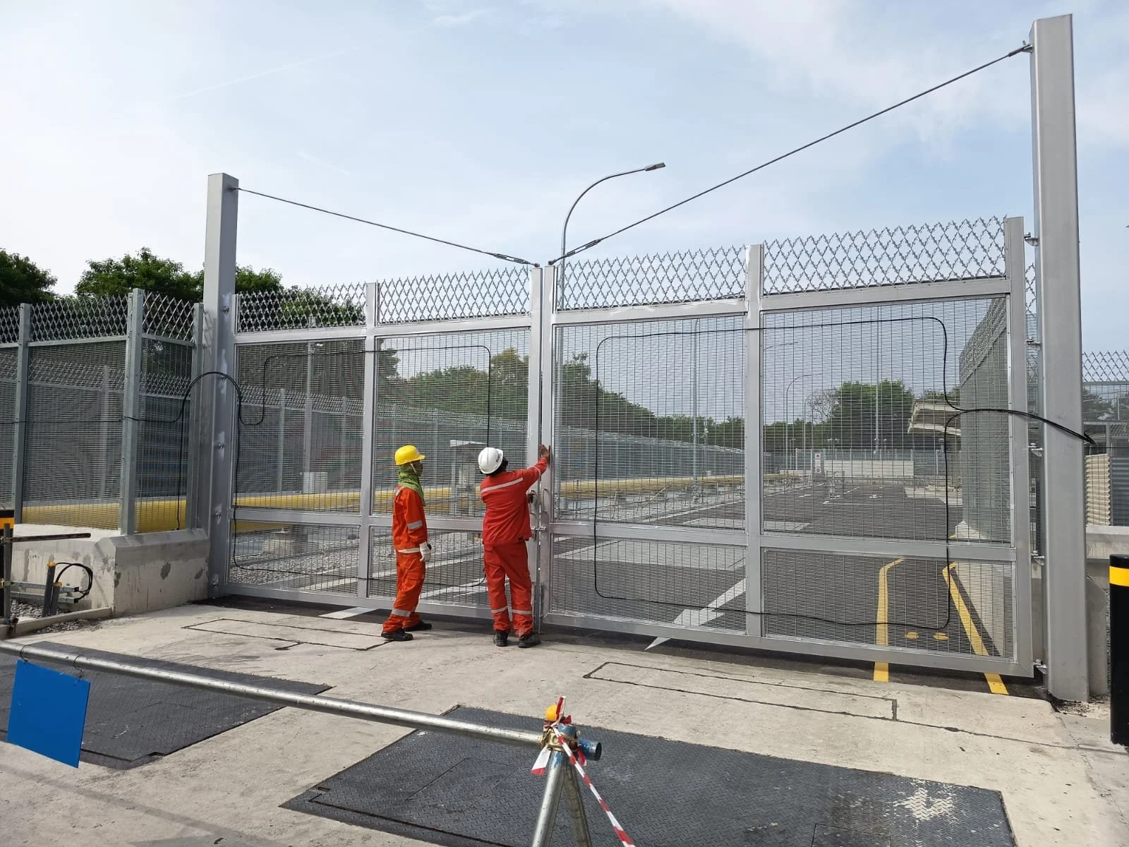 Two construction workers in safety gear working on a large metal gate with mesh panels at a construction site. One worker is pointing at the gate while the other observes.