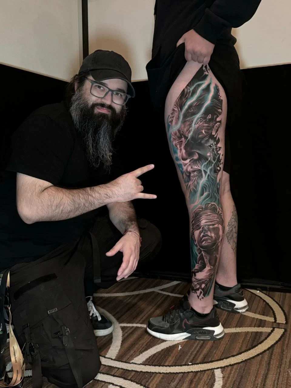 A man with a beard and glasses squatting and making a rock sign with his hand next to a person with an elaborate full-leg tattoo depicting a fantasy scene.