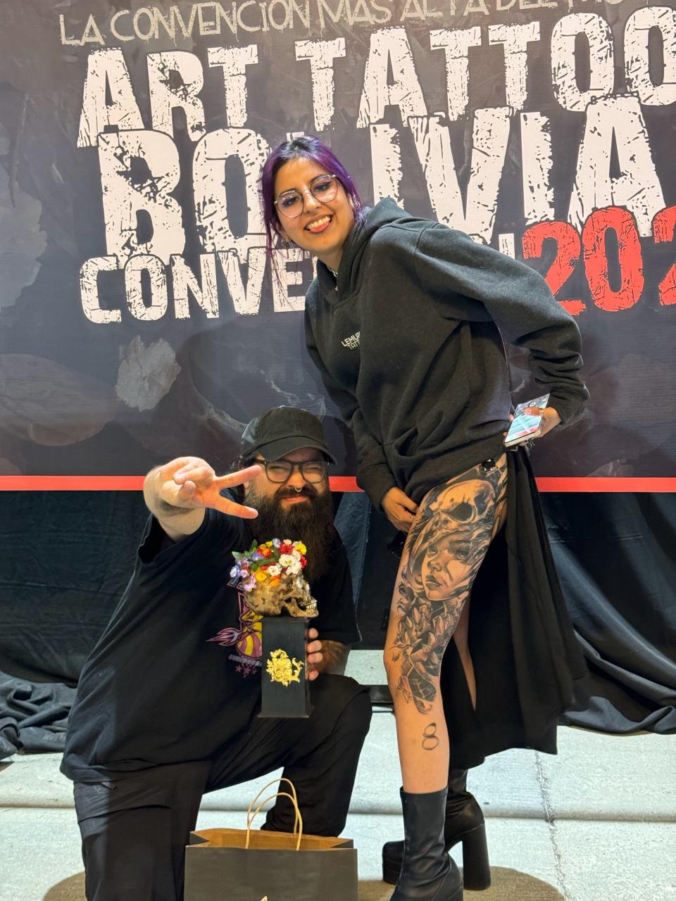 A woman with purple hair, glasses, and a black hoodie standing next to a bearded man wearing glasses and a black cap; the man is squatting and holding a flower arrangement with a skull base. The woman is smiling, holding a phone, and revealing a large tattoo of a woman's face on her thigh with black boots. They are at an art tattoo convention, with a large banner in the background reading "Art Tattoo" and other text.