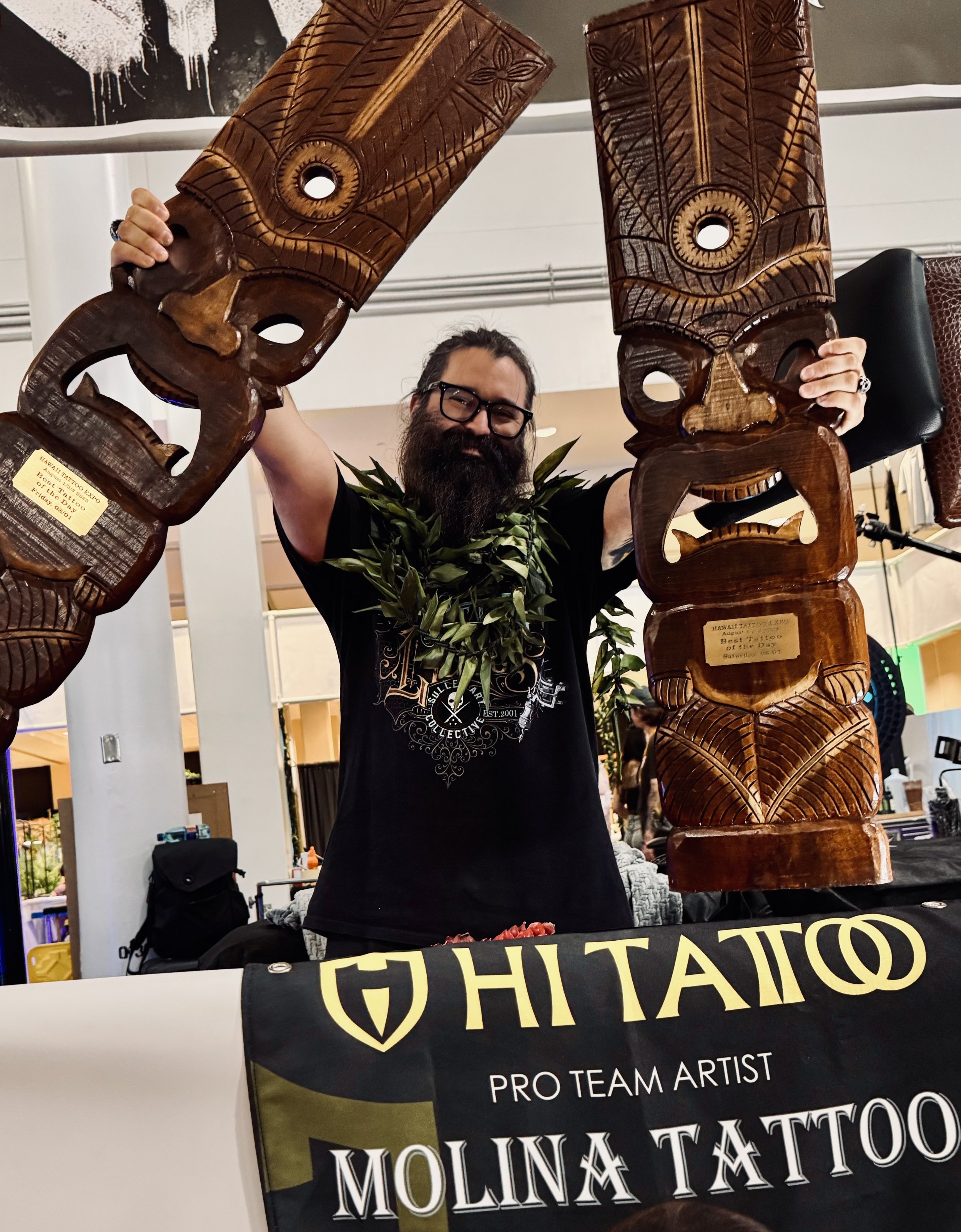 A man with glasses and a beard, wearing a black shirt and a green leaf necklace, is holding two tall wooden tiki statues with carved faces and patterns, standing behind a table with a black cloth that reads 'HIT TATTOO PRO TEAM ARTIST MOLINA TATTOO'. The man is smiling.