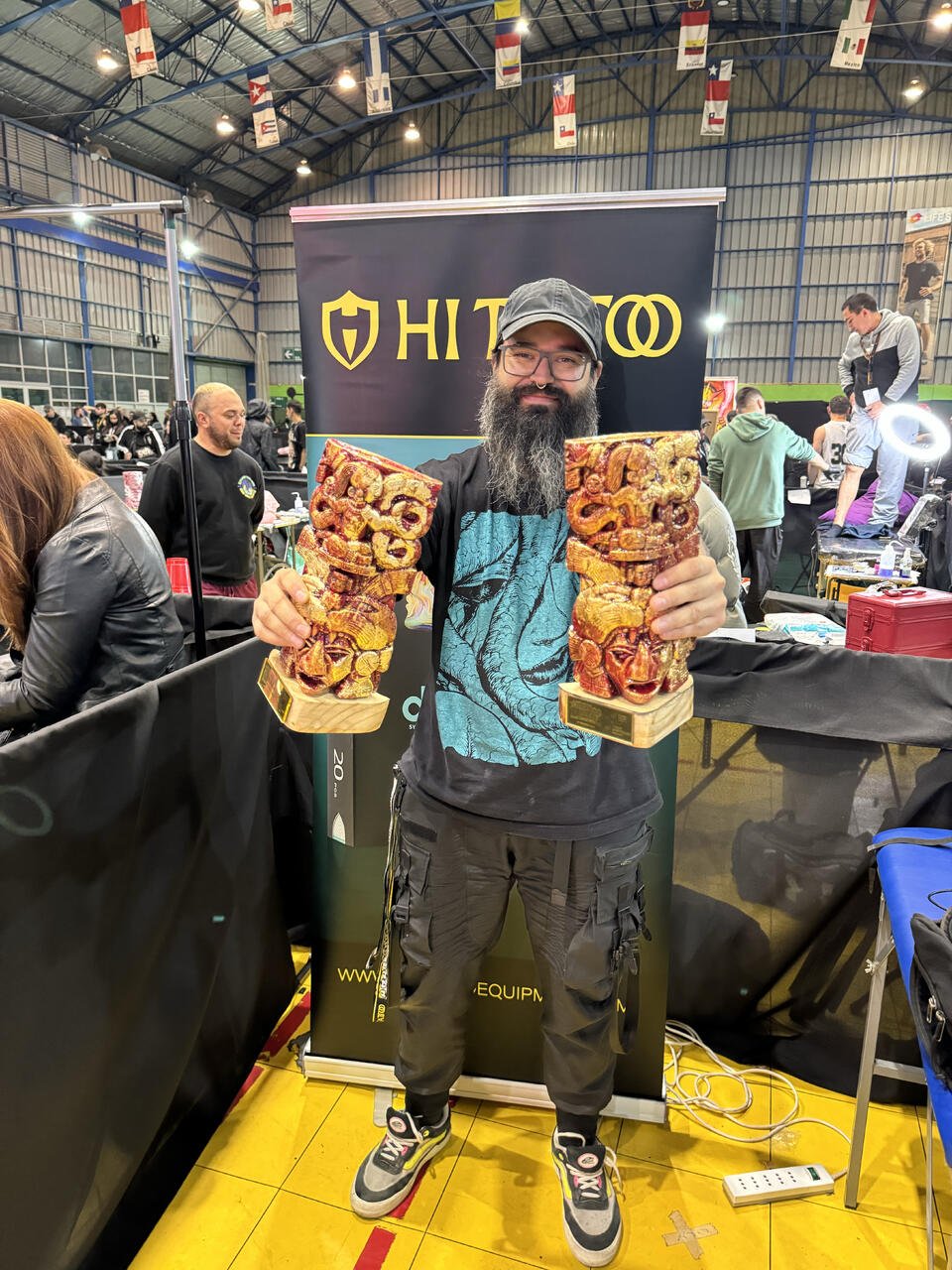 A man with a beard, glasses, and a cap holding two carved tiki statues at an indoor event or convention.