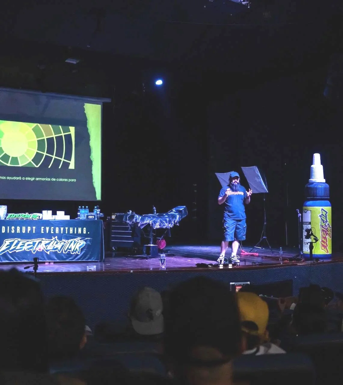 A man on stage giving a presentation in a dark room with a large screen, a table with water bottles, and a giant bottle of glue as a prop.