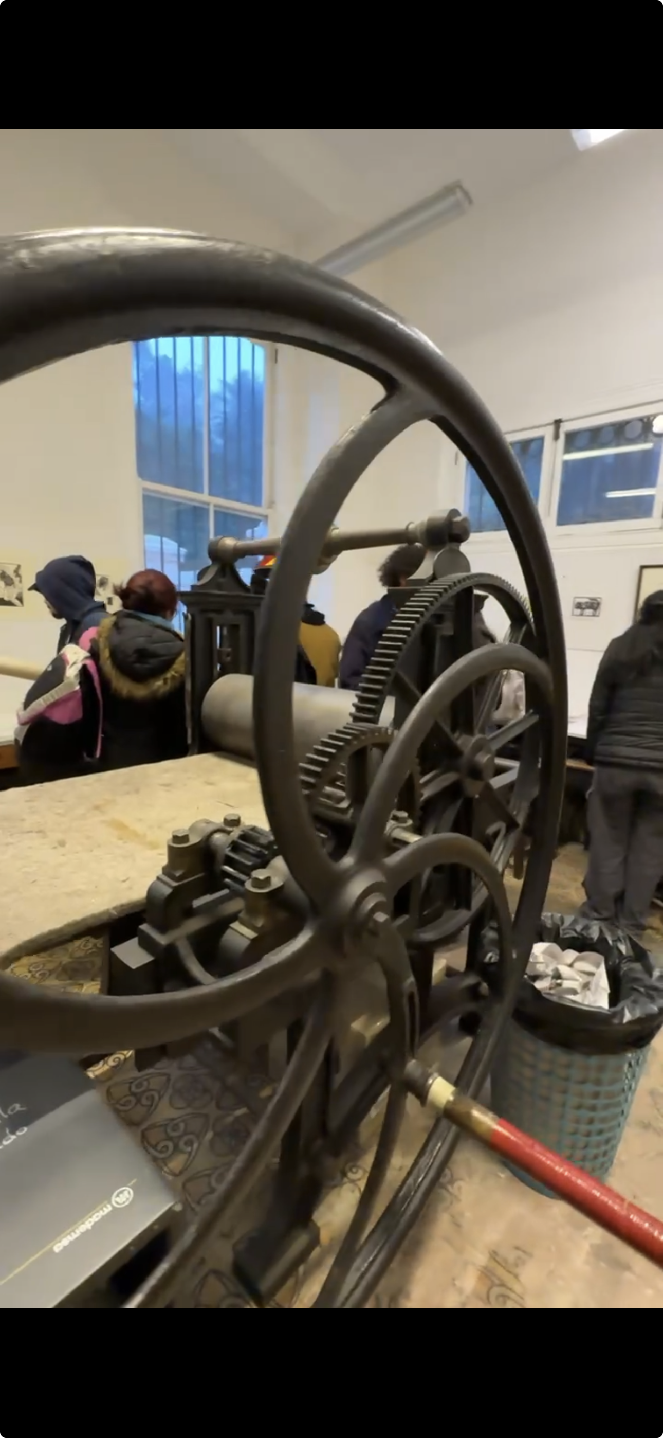 View of a vintage printing press or engraving machine in a museum or exhibit, with people observing in the background.