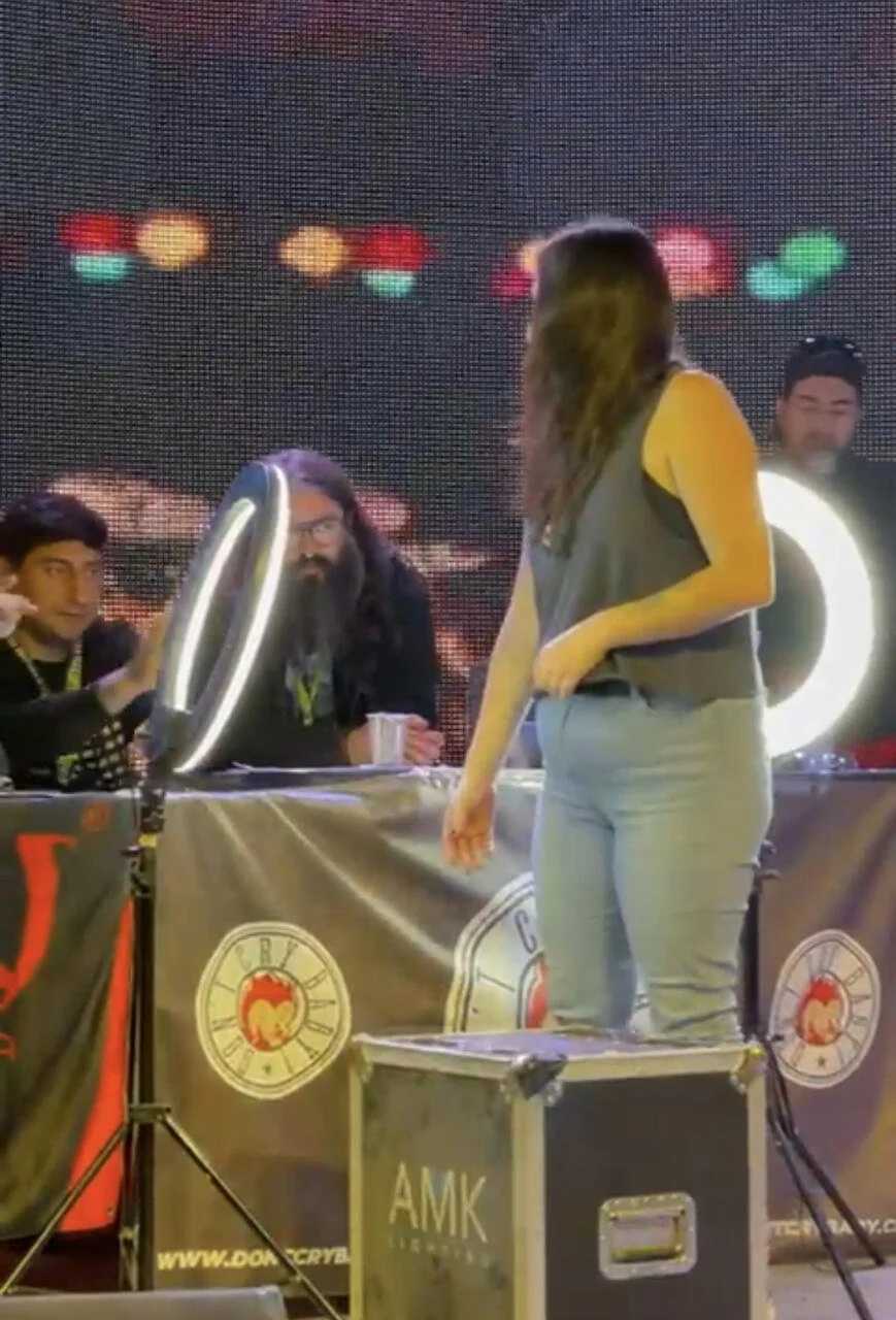 A woman standing on a flight case, holding a ball, facing a panel of judges at a televised event. The judges are seated behind a table with a banner and are looking at her; one of them is wearing glasses and has long hair and a beard.