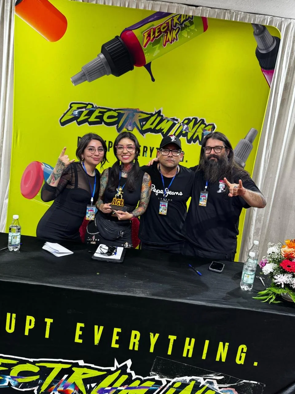Four people standing behind a black table at a convention or event, with a bright yellow backdrop advertising 'Electrum Ink' and tattoo supplies. The group is smiling and making hand gestures, with one person holding a black and yellow 'AFC' sign, framed by lace and flowers on the table.