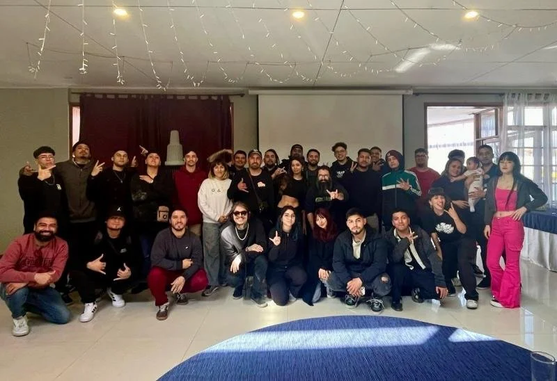 Group of people posing indoors, some making hand gestures, with a projector screen and windows in the background.
