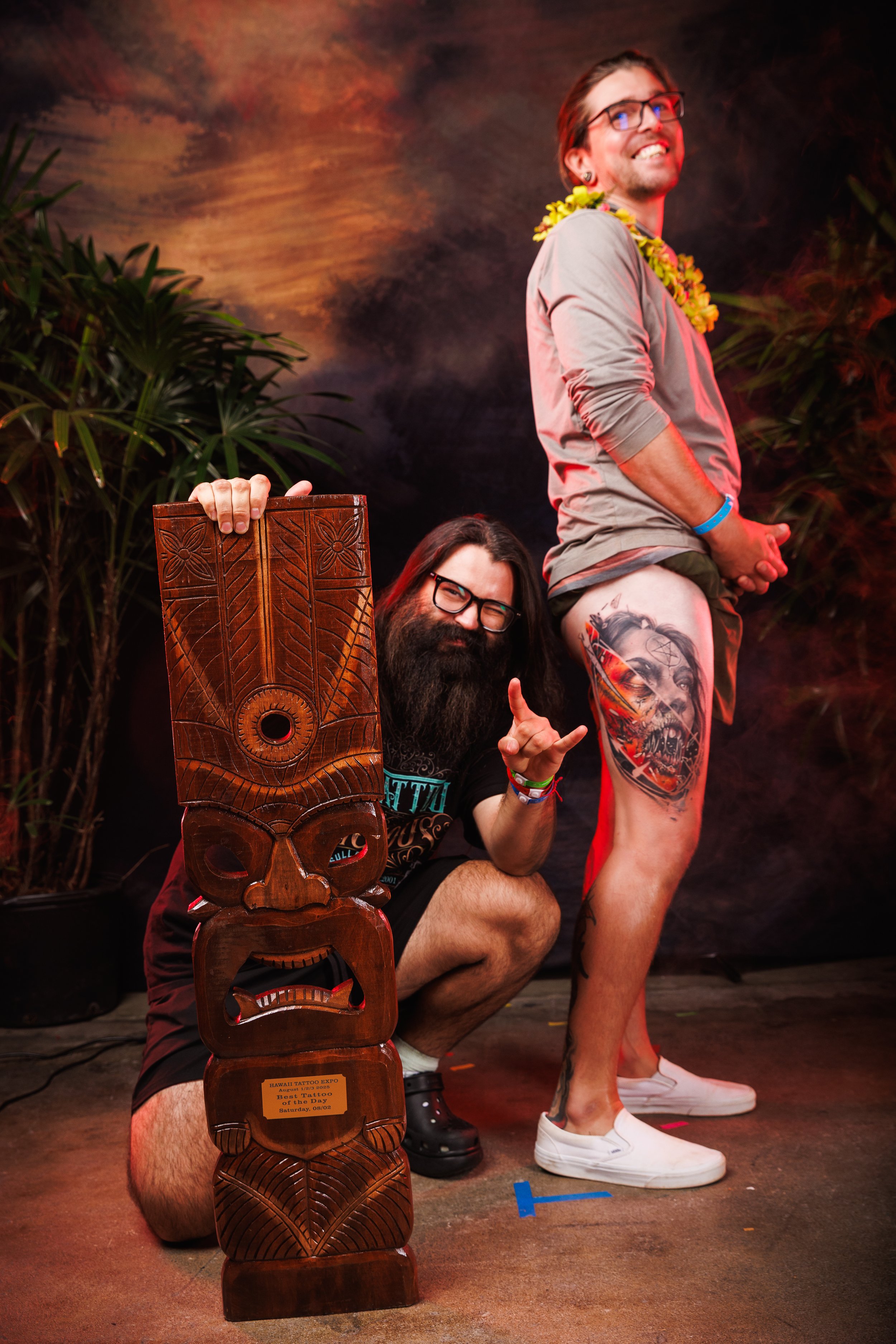 Two men at a tattoo expo, one kneeling beside a tiki sculpture and the other standing showing off a large tattoo on his thigh. The standing man has a floral lei, glasses, and is smiling. The seated man has a beard, glasses, and is making a rock and roll hand gesture.