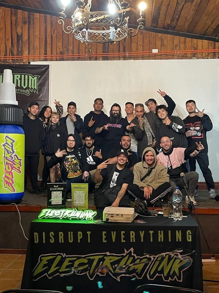 Group of people posing on a stage at an event called Electrum Ink, with a banner that says 'Disrupt Everything.' A large glue bottle prop and QR code signs are on a table in front. The setting has wooden walls and a chandelier hanging from the ceiling.