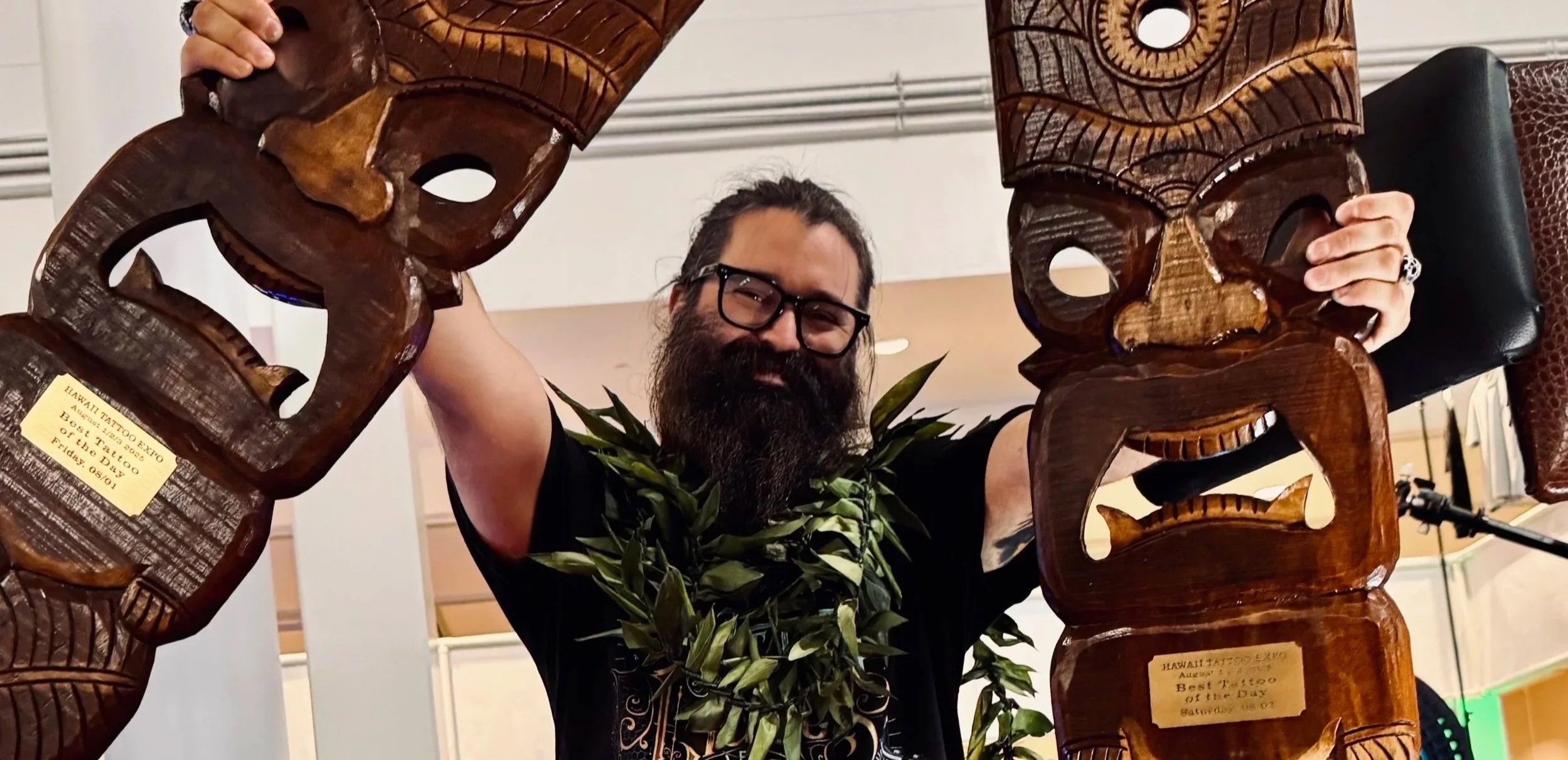 Man with glasses and a beard wearing a black T-shirt, holding two carved wooden tiki masks, standing behind a green leafy plant, in a room with a white wall.