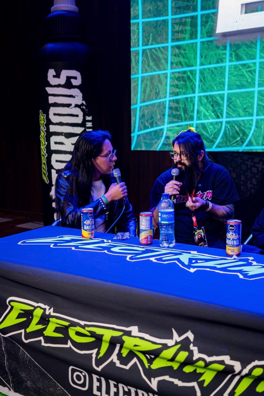 Two people speaking into microphones seated at a table with drinks, behind them a large screen and a themed tablecloth with the text 'ELECTRUM'.