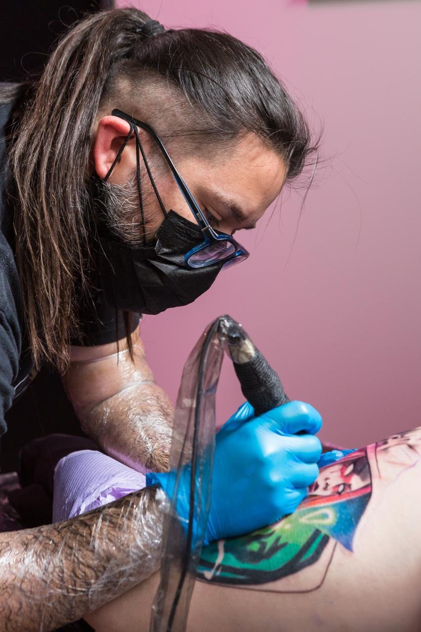 Tattoo artist wearing glasses, face mask, and gloves tattooing a colorful design on a person's forearm.