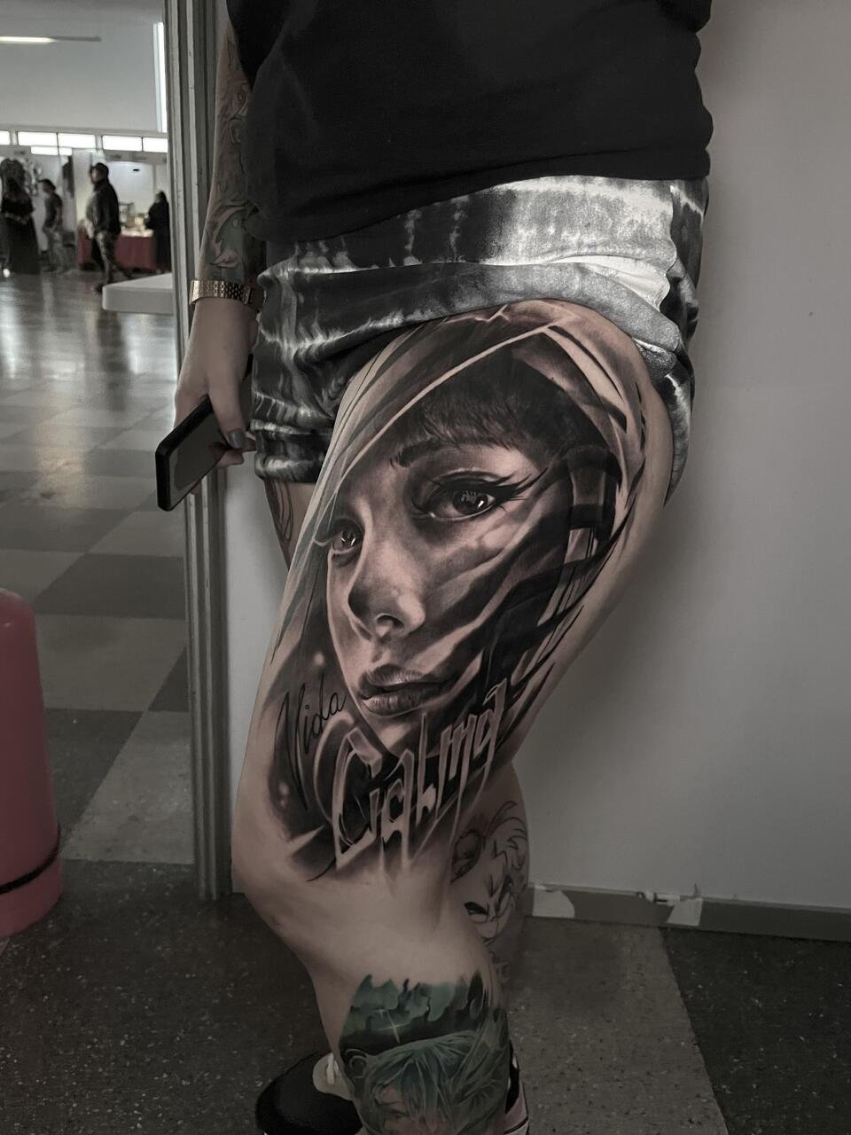 Tattooed leg with detailed black and gray portrait of a woman's face, surrounded by stylized text and additional tattoo art, in an indoor setting.