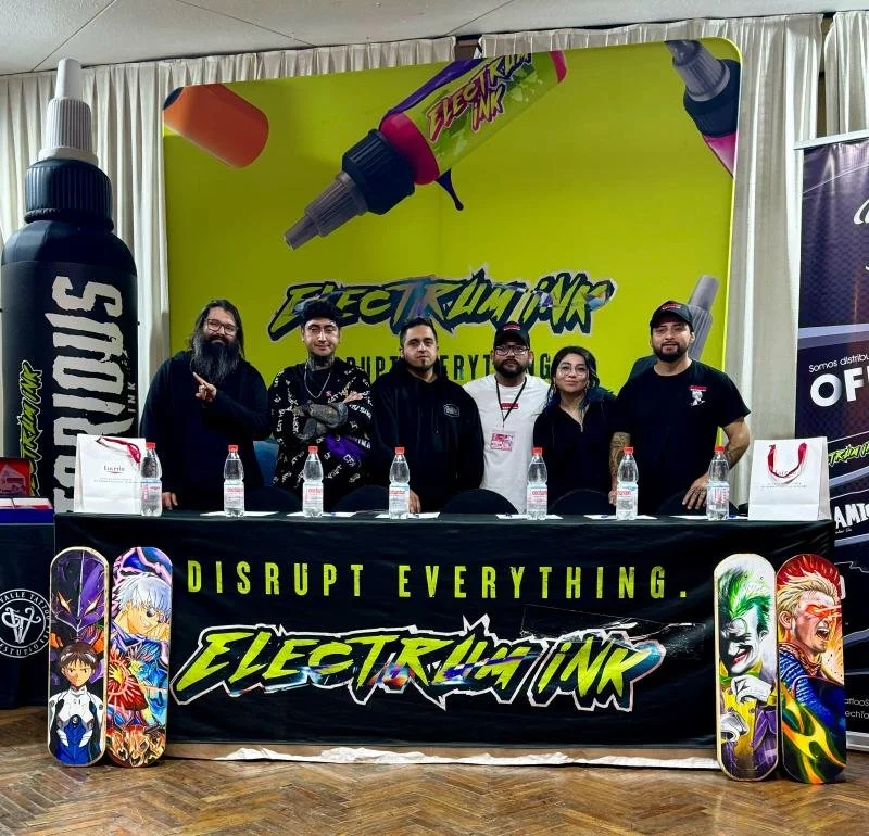 People standing behind a table with bottles in front, at an event booth for Electric Ink, a brand of spray ink or spray paint, with anime-themed skateboard designs displayed at the sides, and a large spray bottle graphic on the backdrop.