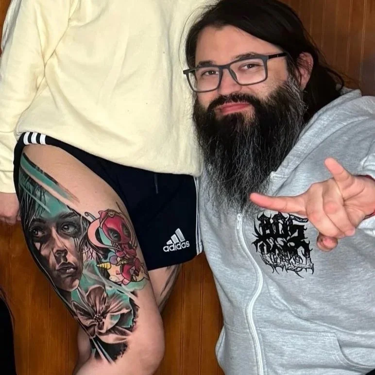 A man with long dark hair, glasses, and a beard is smiling and making a hand gesture. The photo shows his upper body and part of another person's leg, who has a tattooed thigh with colorful, detailed tattoos. The tattoos include a woman's face, a cartoonish character, and flowers.