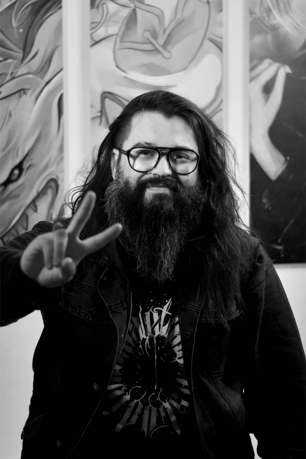 A man with long hair and a beard wearing glasses, making a peace sign gesture with his right hand, standing in front of artwork with abstract and cartoon-like designs, in a black and white photograph.