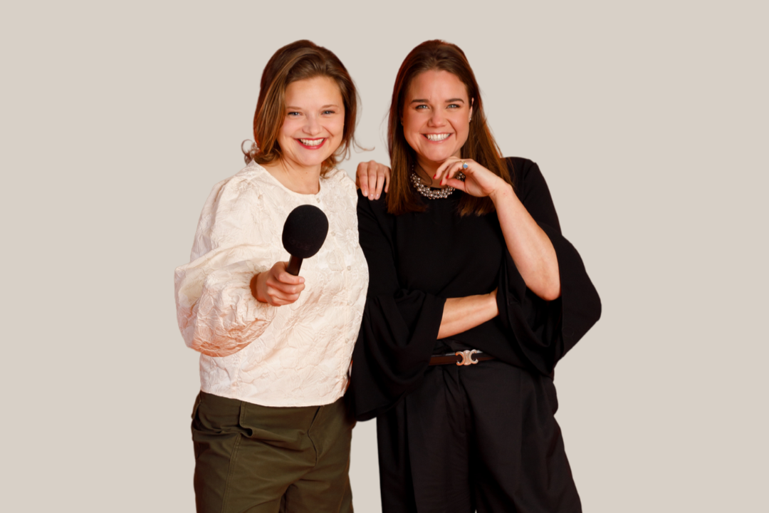 Two smiling women posing against a plain orange background. One has shoulder-length brown hair, wears a black blazer, and a patterned scarf. The other has her blonde hair in a bun and wears a colorful floral blouse.