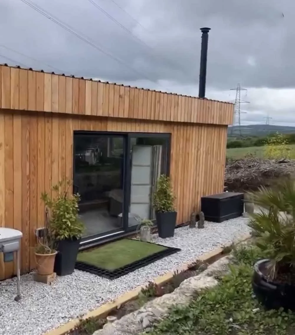Exterior of a small wooden house with sliding glass door, potted plants, and outdoor gravel area, with cloudy sky and power lines in the background.