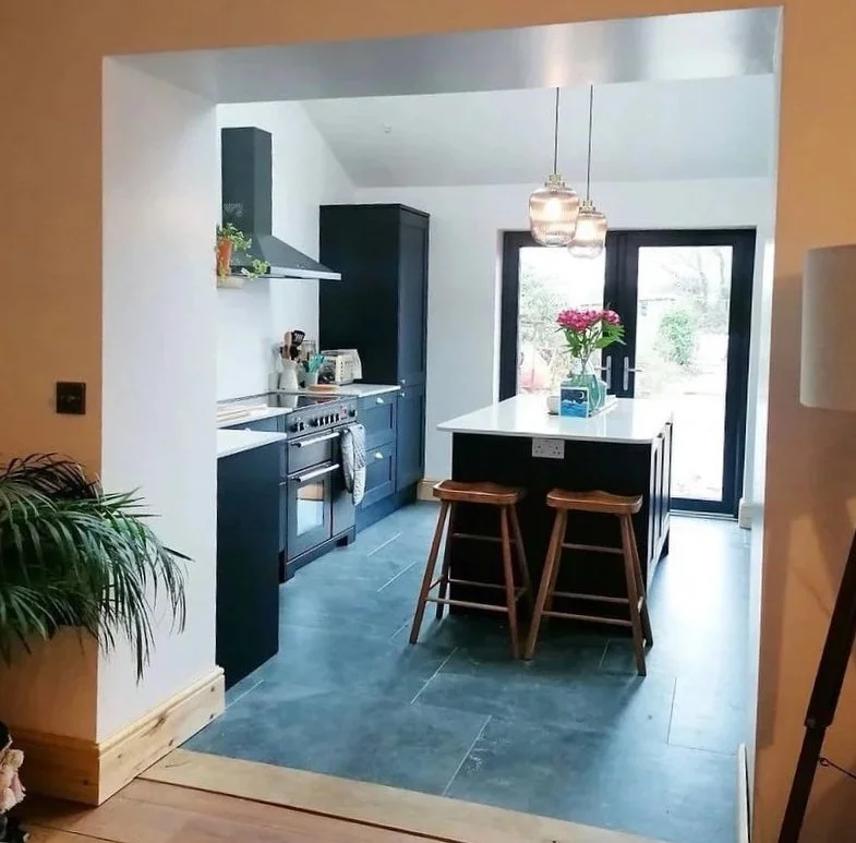 A modern kitchen extension with dark blue cabinets, a white kitchen island with a dark base, and bar stools. Hanging pendant lights and large french doors leading outside. The kitchen is visible from the original room.