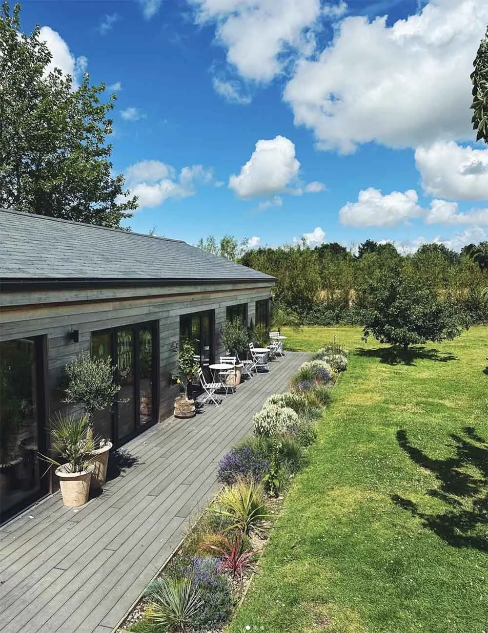 St Kew Farm shop building, A wooden deck lines the outside of the wood clad commercial building with black-framed glass doors, potted plants, and outdoor seating, surrounded by a grassy lawn with trees and a blue sky with white clouds.