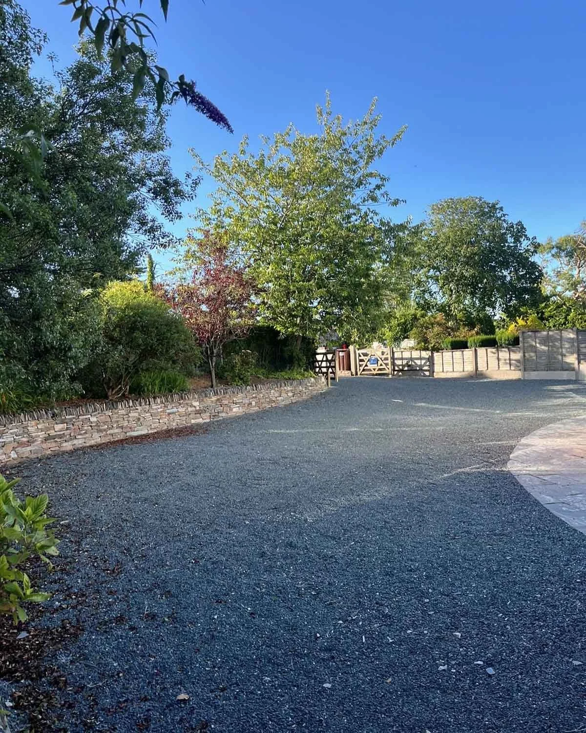 A newly landscaped gravel driveway with a curved edge, bordered by a low stone wall and surrounded by trees and shrubs under a blue sky.
