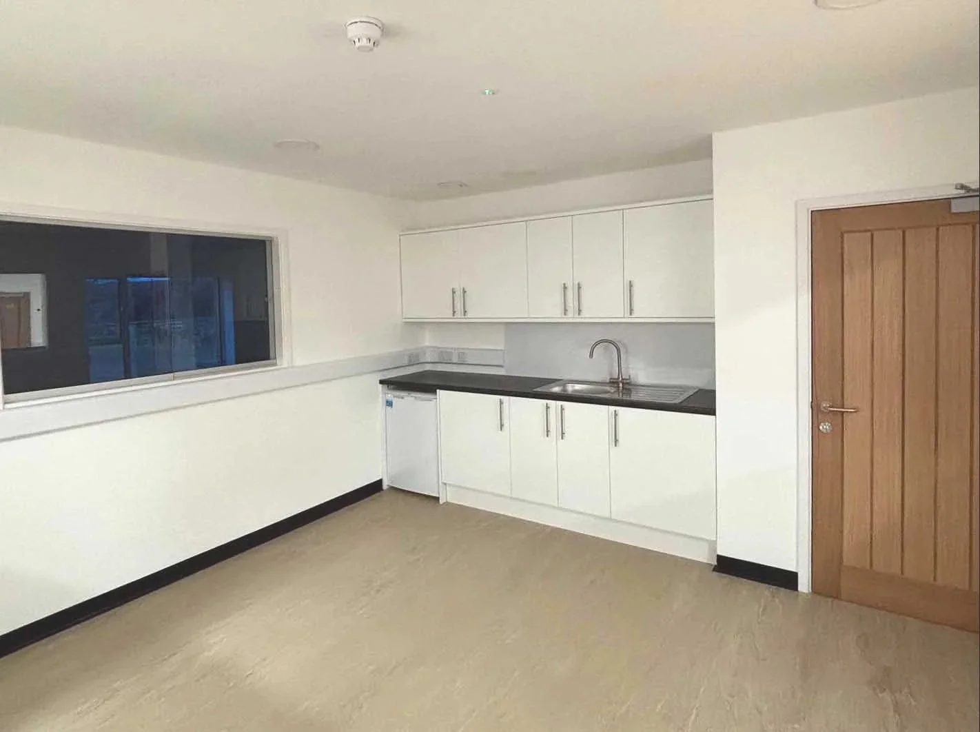 Inside St Tudy Community center the large kitchen with white cabinets, a black countertop, a sink, a small refrigerator, a large window is sat on a practical and easy clean flooring that goes throughout the building.