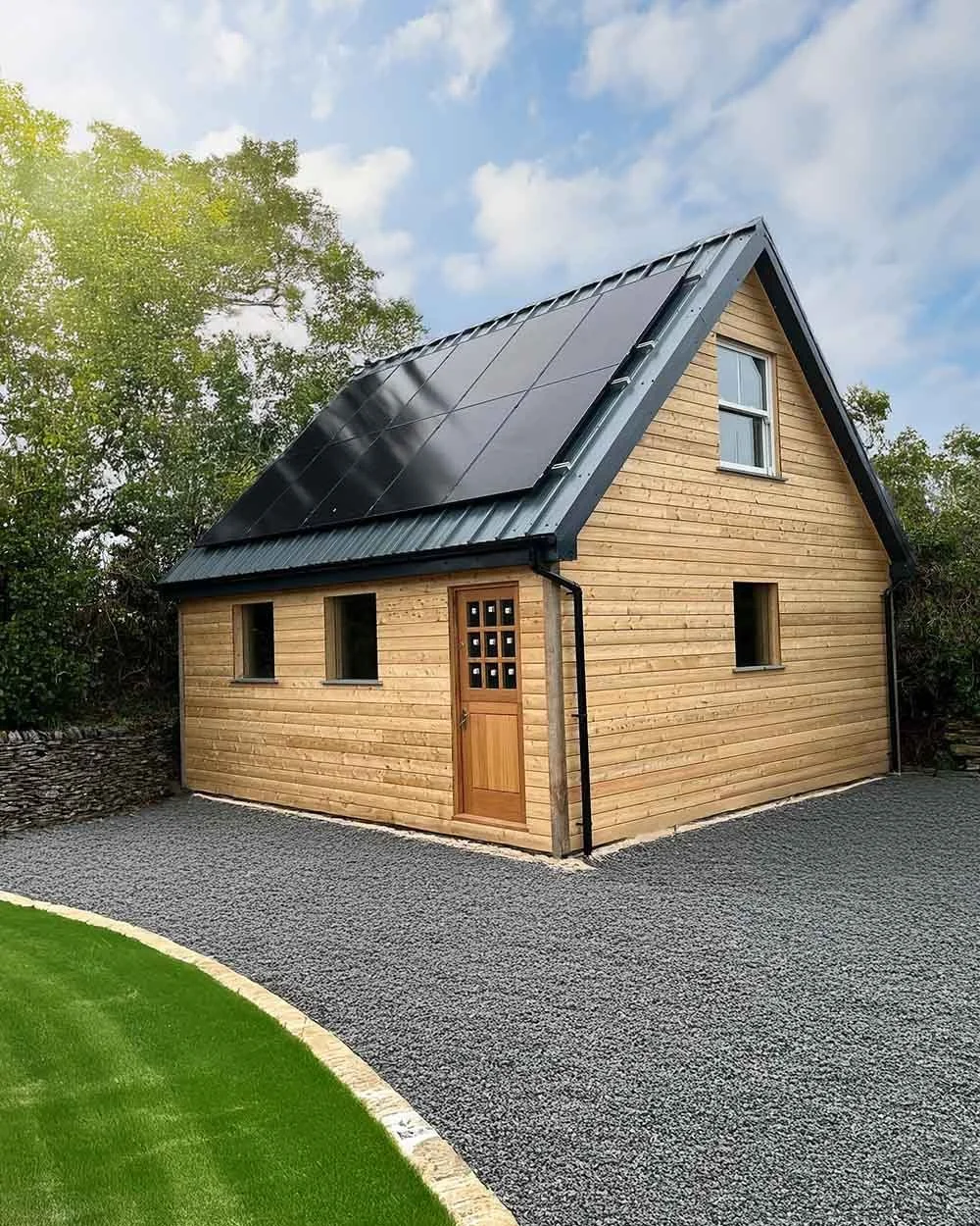 A small wooden house built by Runnals Construction in Cornwall with a sharply pitched metal roof featuring solar panels. The house has three small rectangular windows and a wooden door with glass panes.