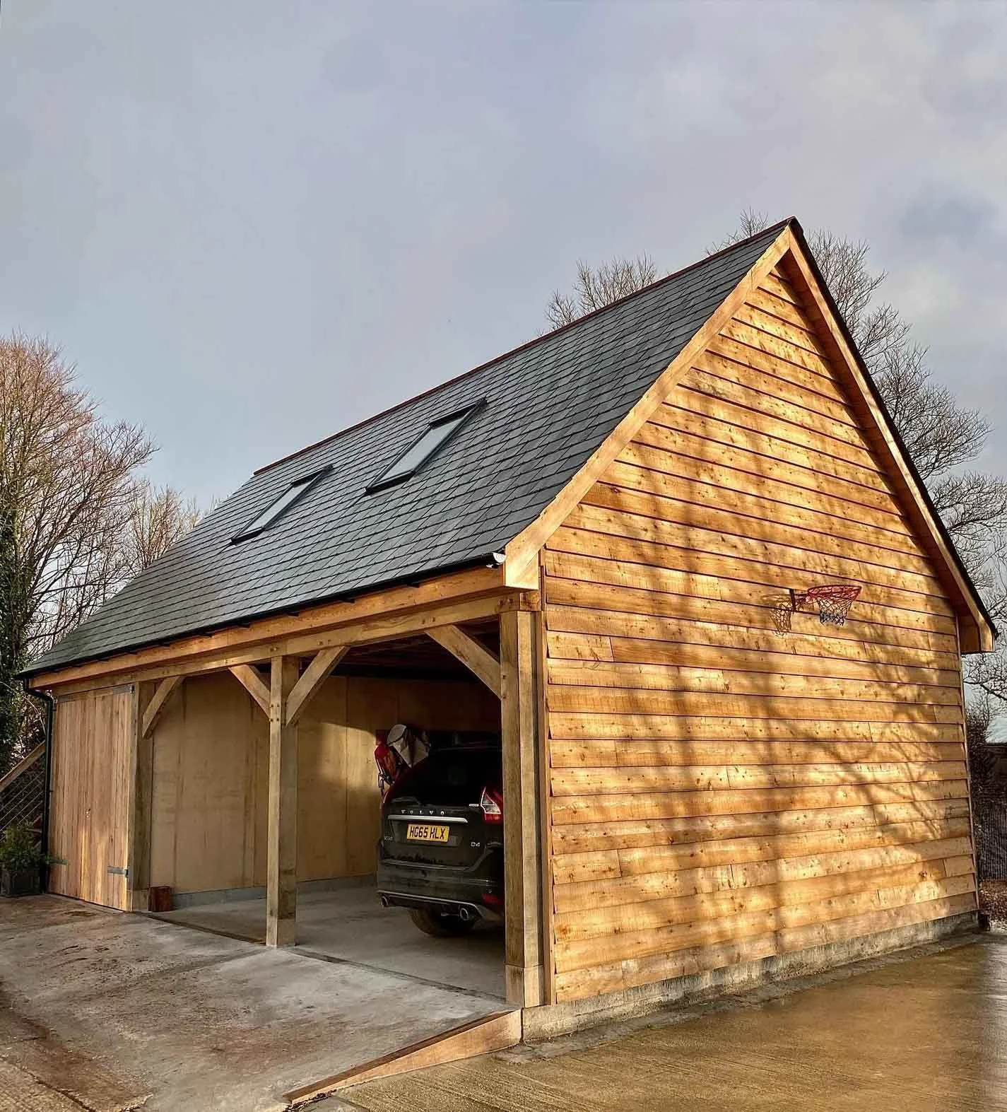 A oak frame garage and two car port . The garage has vertical wooden siding, a sloped roof with two skylights, the upstairs is reach via external wooden steps and is a large living space.