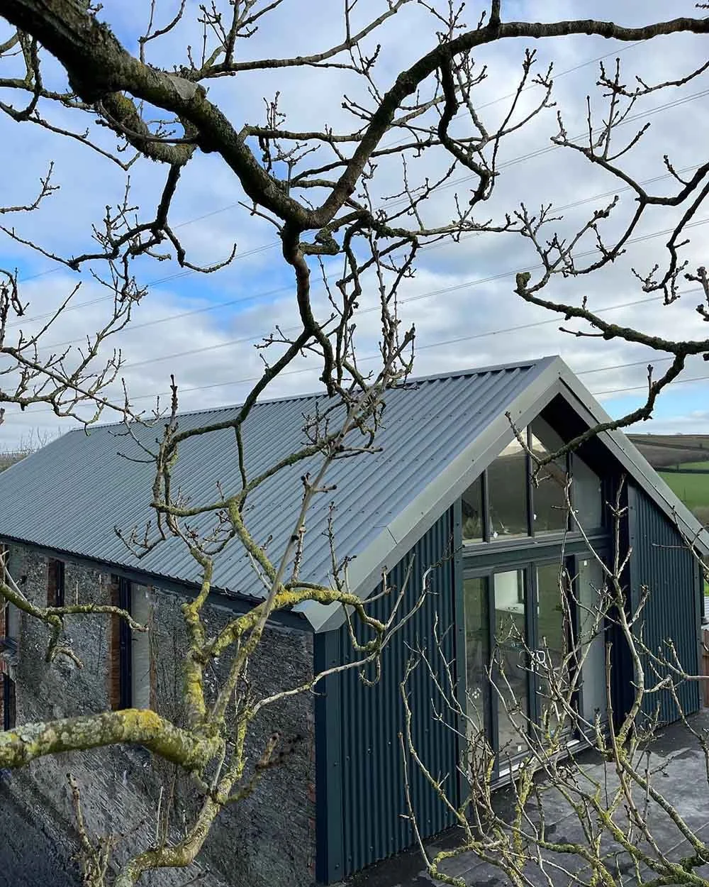 Cornish Barn Converstion with zinc roof and large two story windows.  Side wall is original stone, front of building is corregated grey metal.  Old trees in the foreground has not been distrubed during build