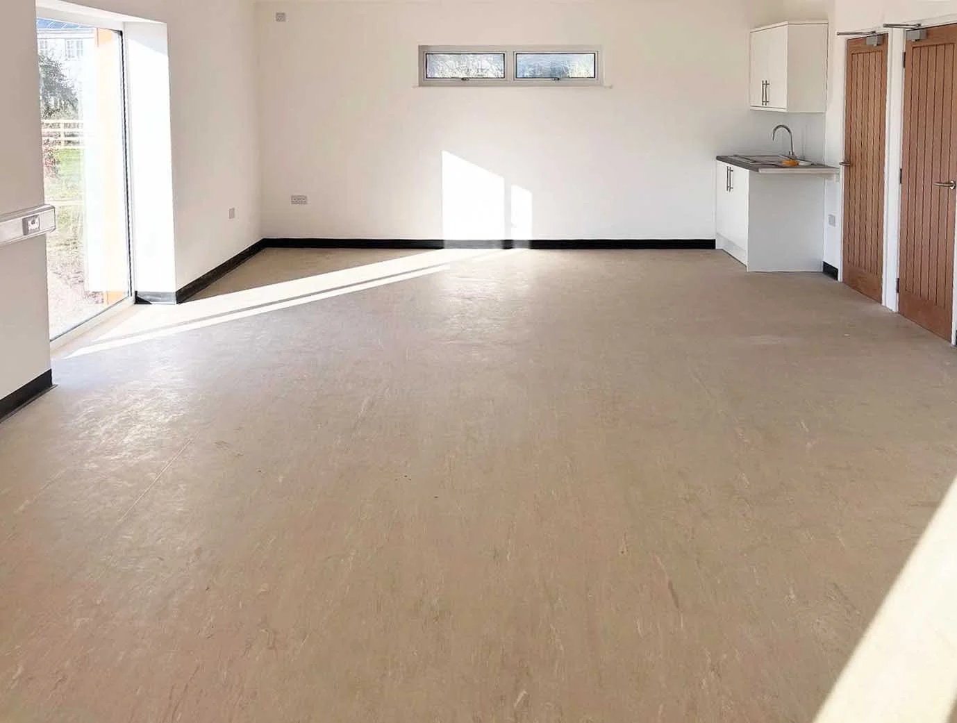 Communal area at St Tudy Community center with practical flooring, white walls, a small kitchen area with white cabinets, a sink, and a wooden double door.