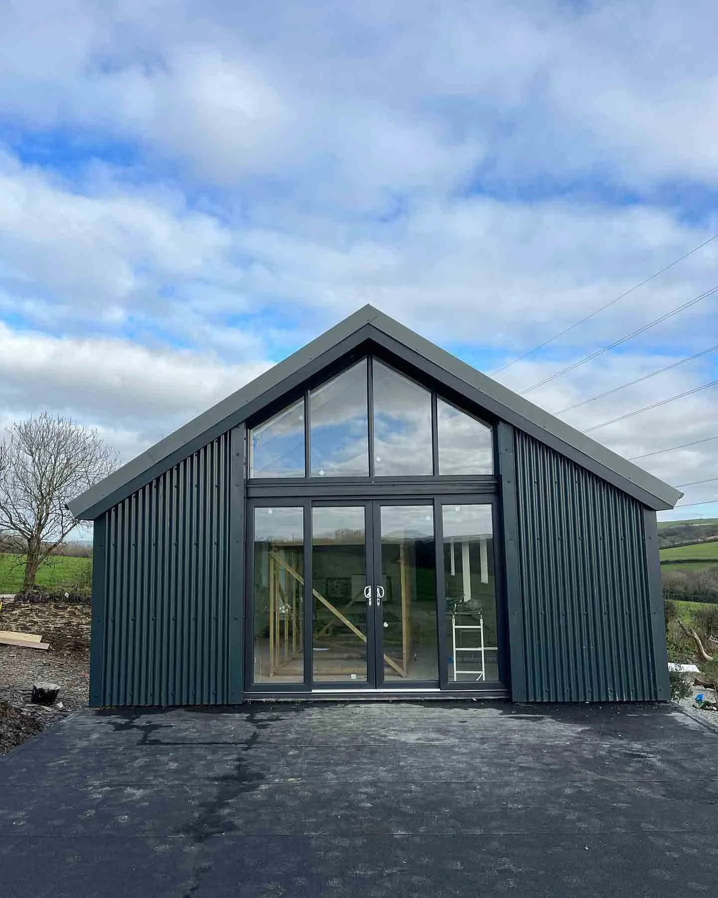 A black metal building with a gable roof and large glass doors and windows, set against a rural landscape with green fields and a tree, under a cloudy sky.  This is the outcome of a barn conversion in Cornwall converted by Runnalls Construction