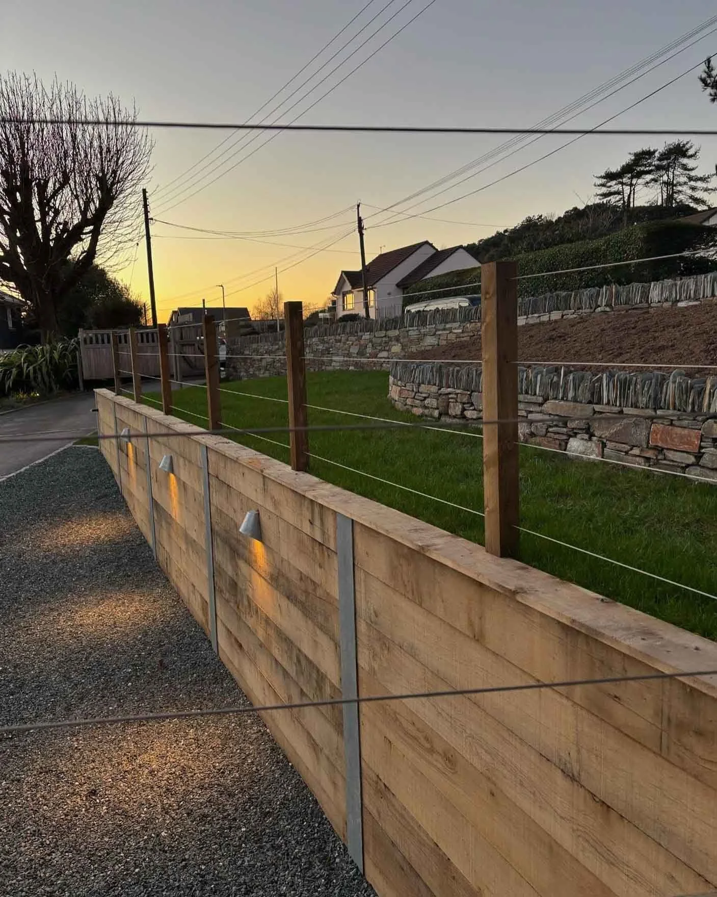 A wooden fence with wire strands and exterior lighting along a gravel driveway at sunset. Behind is a fresh laid lawn and stunning Cornish stone retaining walls creating a boundary for the property and a flower bed ready for planting.