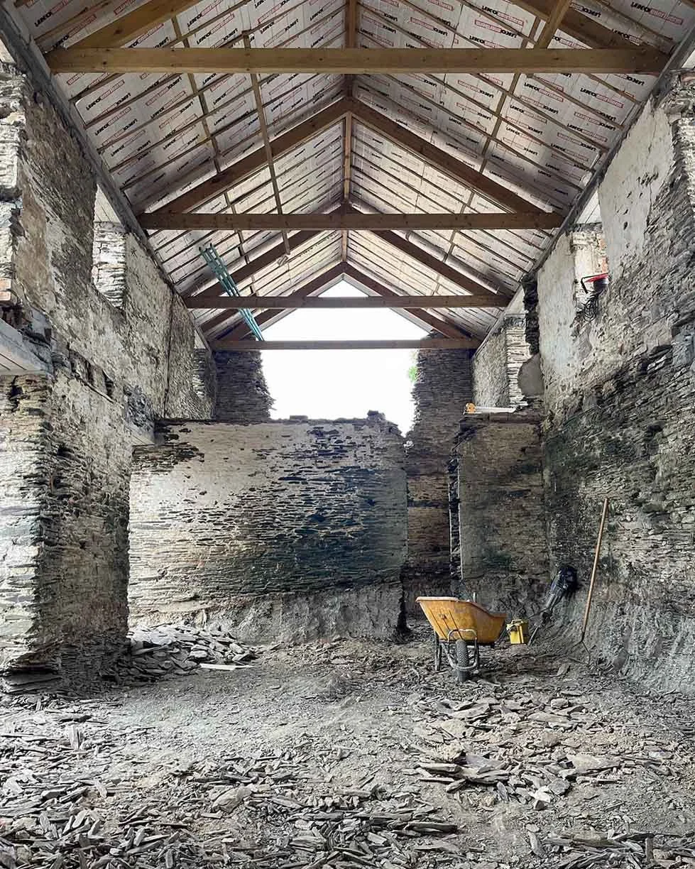 The interior of a historic stone barn under renovation, with exposed stone walls, a wooden roof structure with insulation, and construction tools including a wheelbarrow and shovel.  Transformed to beautiful home in Cornwall by Runnalls Construction